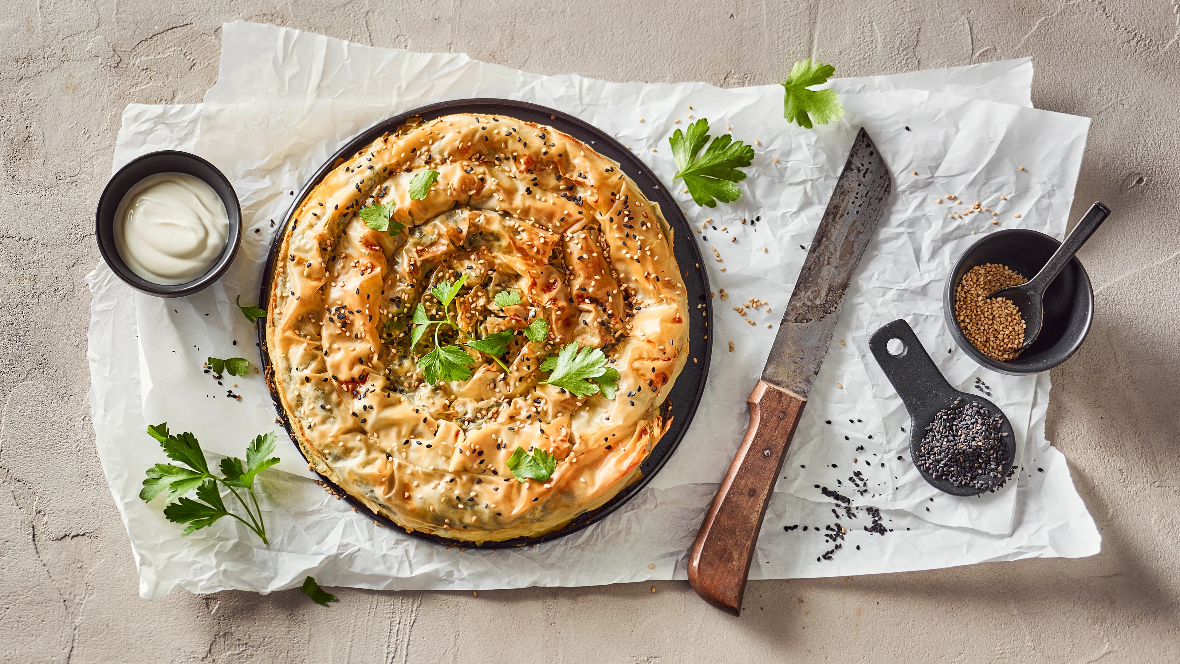 Rezept für türkische Börek-Schnecke Eine knusprig gebackene, gefüllte Teigschnecke, getoppt mit Sesam, auf einem Teller, daneben ein Schälchen mit weißer Creme