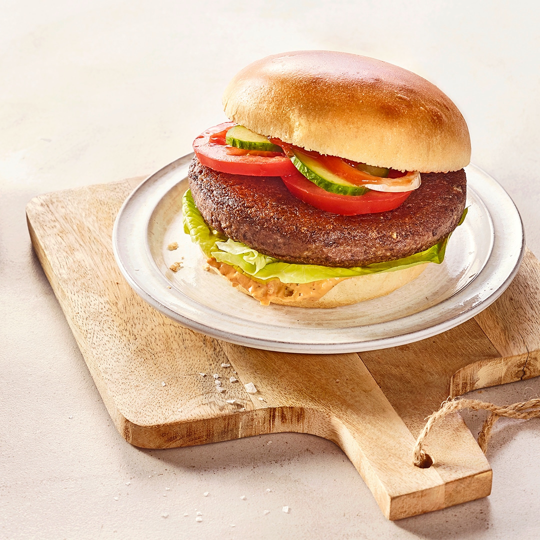 Ein Teller mit Burgerbrötchen, darin braunes Patty, grüne Salatblätter, rote Tomatenscheiben und grüne Gurkenscheiben.