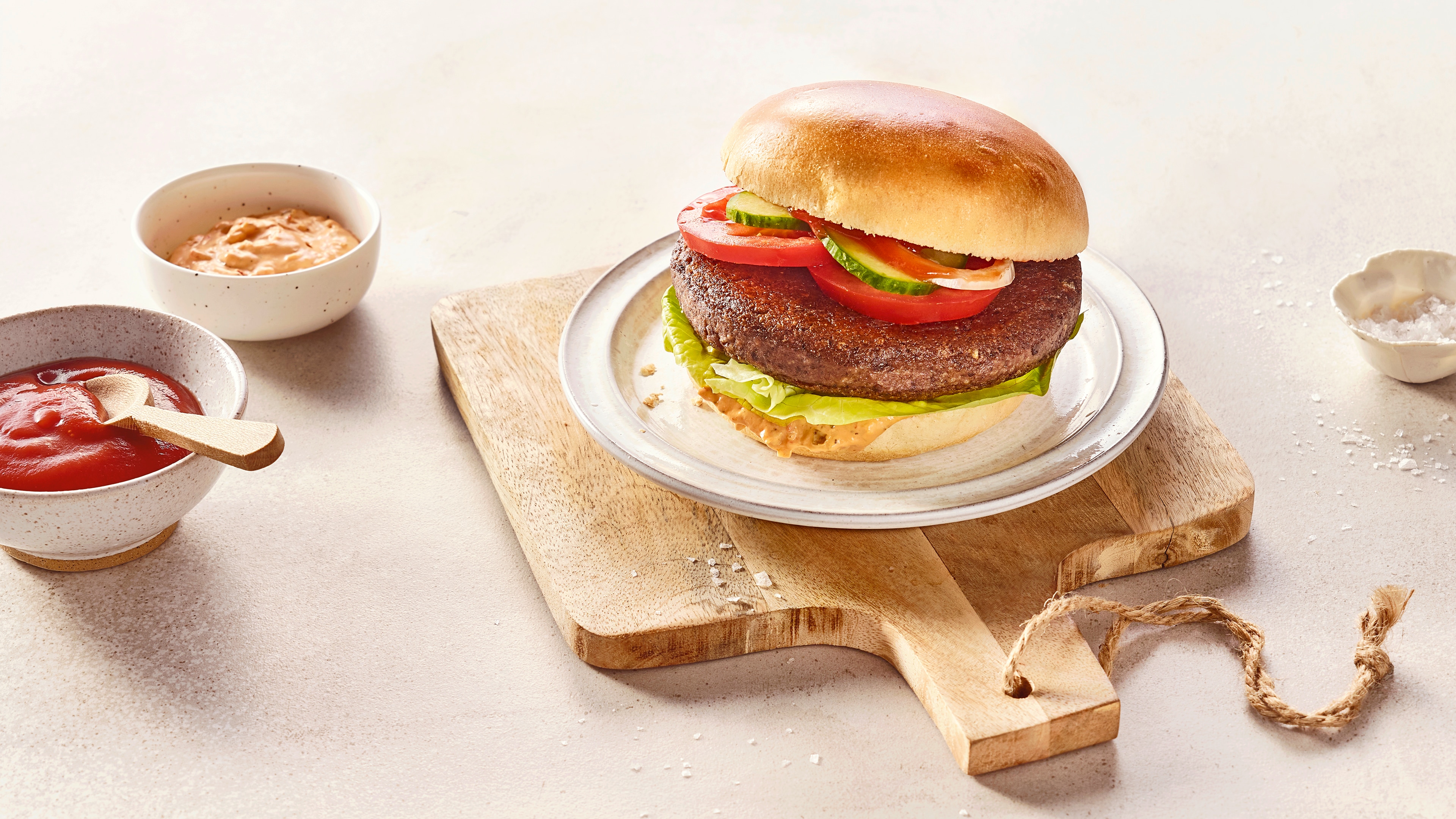 Ein Teller mit Burgerbrötchen, darin braunes Patty, grüne Salatblätter, rote Tomatenscheiben und grüne Gurkenscheiben.