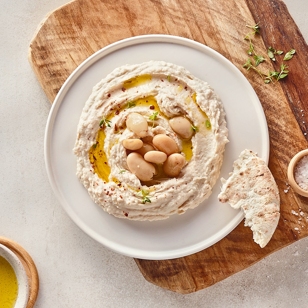 Ein Teller mit einer hellen, dicken Creme, getoppt mit klarem Öl und weißen Bohnen, daneben ein Stück helles Brot