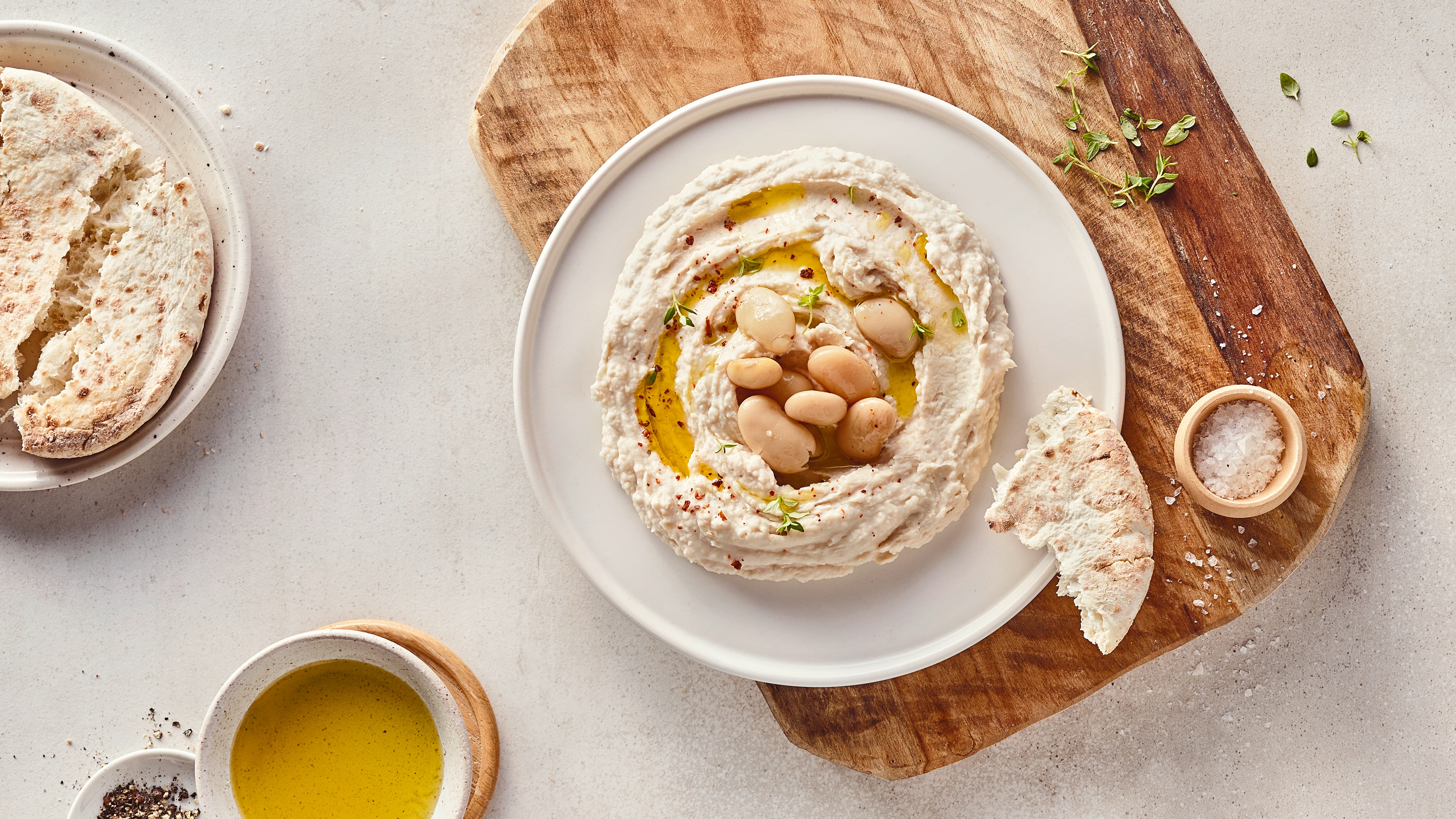 Rezept für Bohnenpüree Ein Teller mit einer hellen, dicken Creme, getoppt mit klarem Öl und weißen Bohnen, daneben ein Stück helles Brot