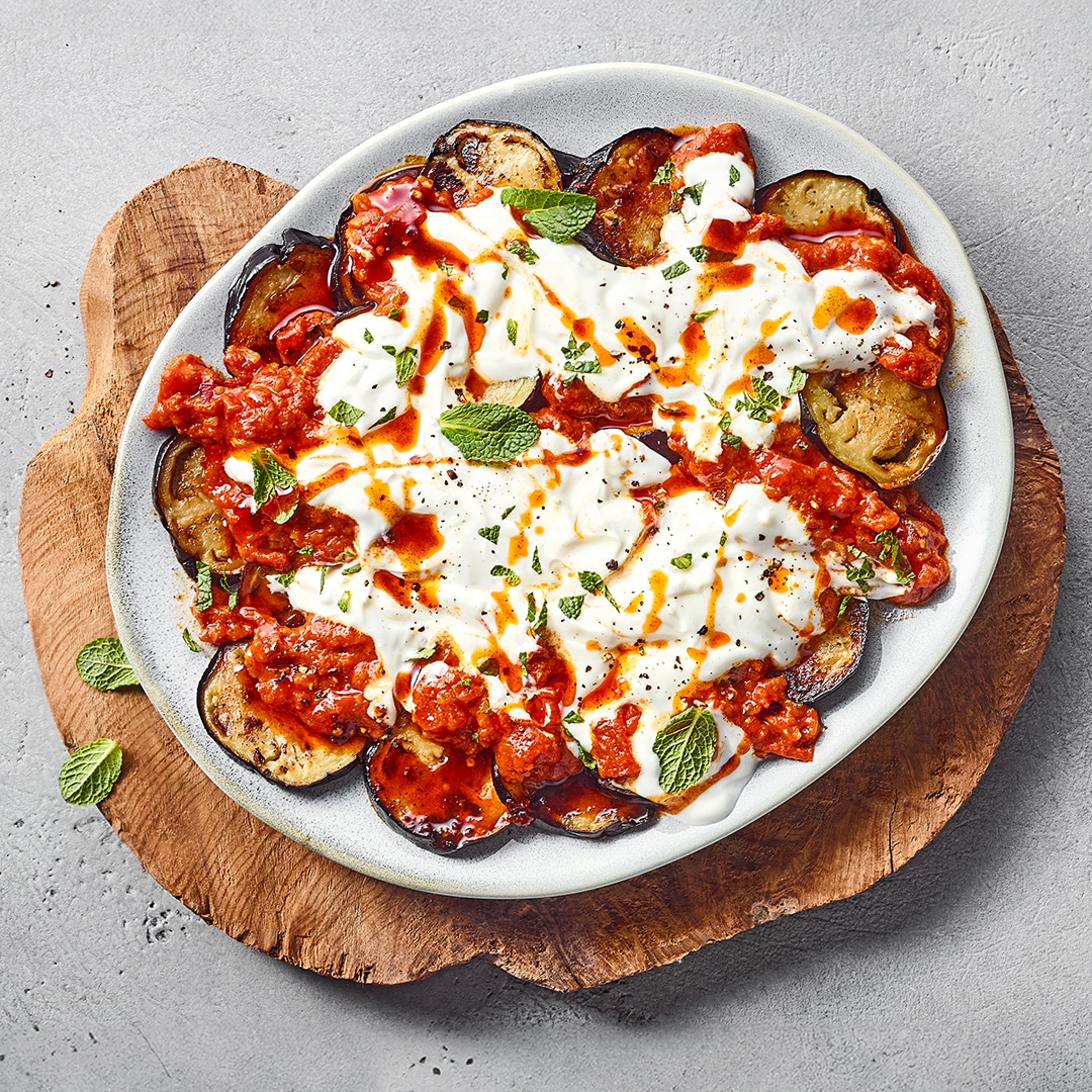 Ein Teller mit gebratenen Auberginenscheiben, roter Tomatensoße, weißem Joghurt und grünen Minzblättern.