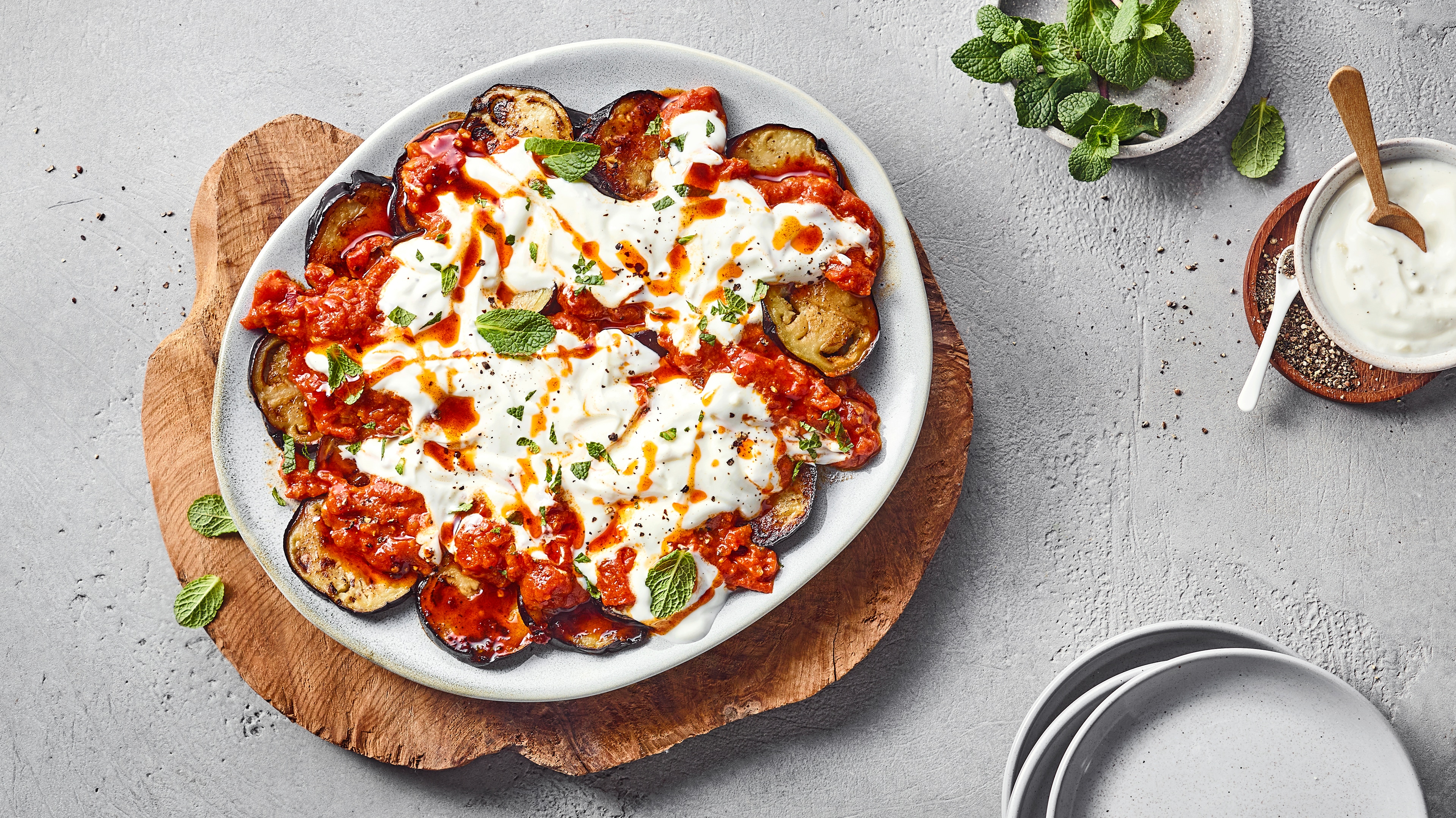 Ein Teller mit gebratenen Auberginenscheiben, roter Tomatensoße, weißem Joghurt und grünen Minzblättern.