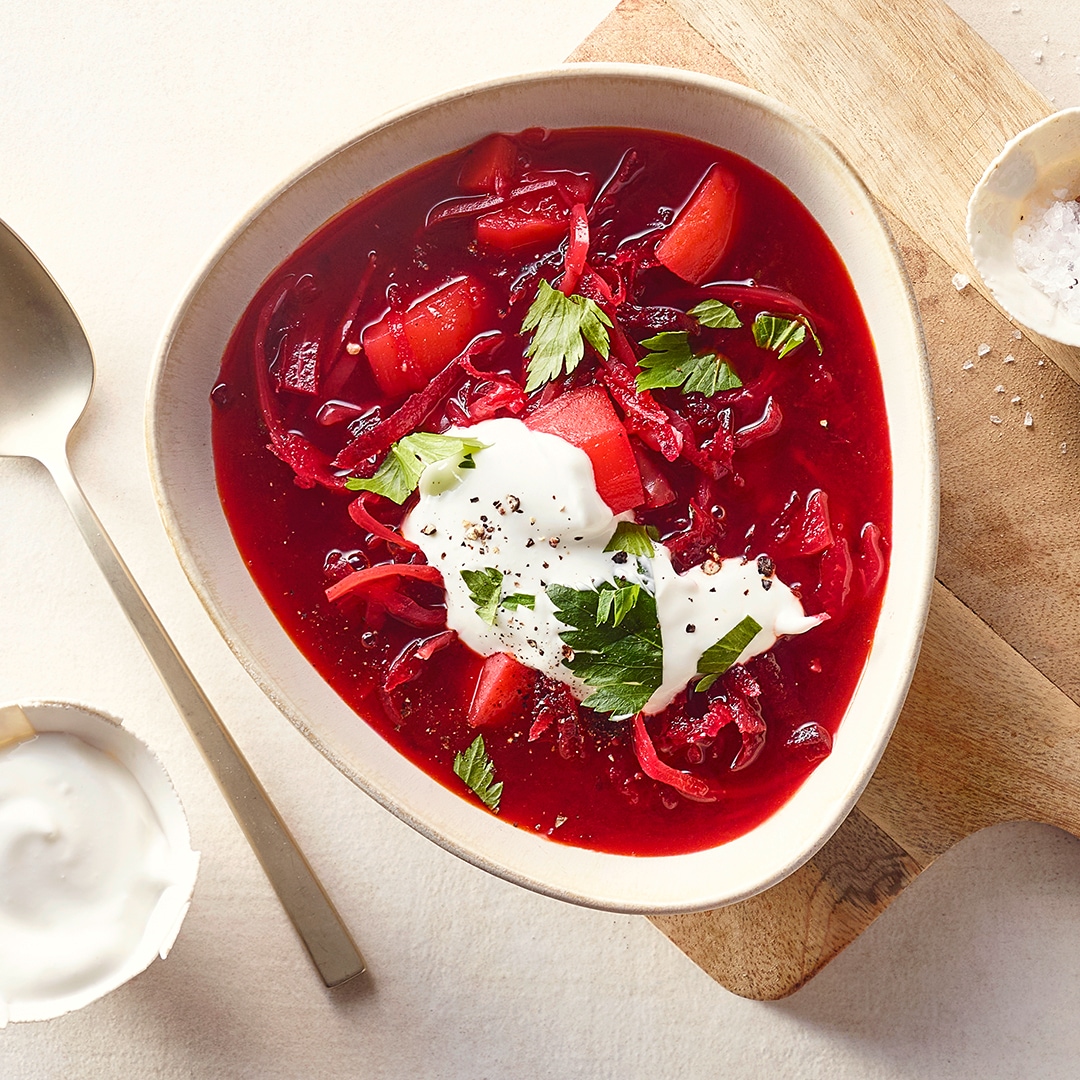Eine Schale mit roter Suppe aus Rote Bete und Gemüse, garniert mit einem Klecks weißer Creme und frischen grünen Kräutern