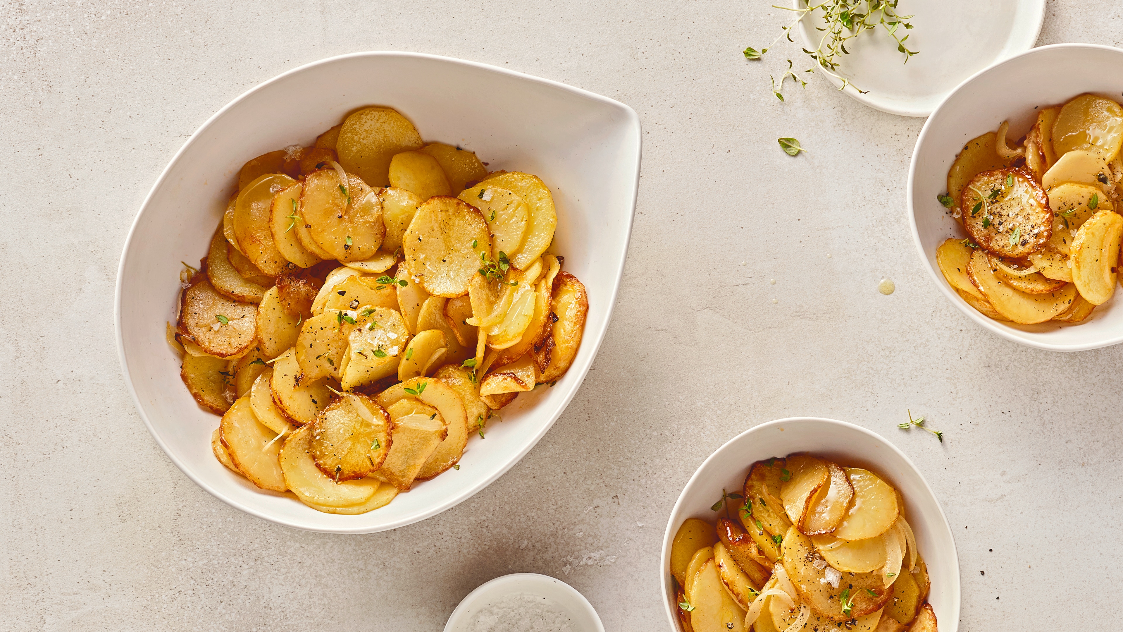 Rezept für Bratkartoffeln aus dem Airfryer Eine tropfenförmige Schale mit dünnen, gelbgoldenen Kartoffelscheiben, garniert mit etwas frischem Thymian