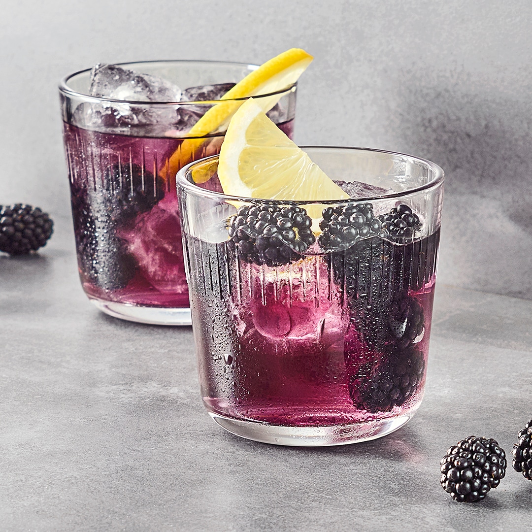 Zwei durchsichtige Gläser mit violettem Eistee, gefüllt mit Brombeeren, Eiswürfeln und Zitronenschalen. Rundherum liegen weitere gefrorene Brombeeren.