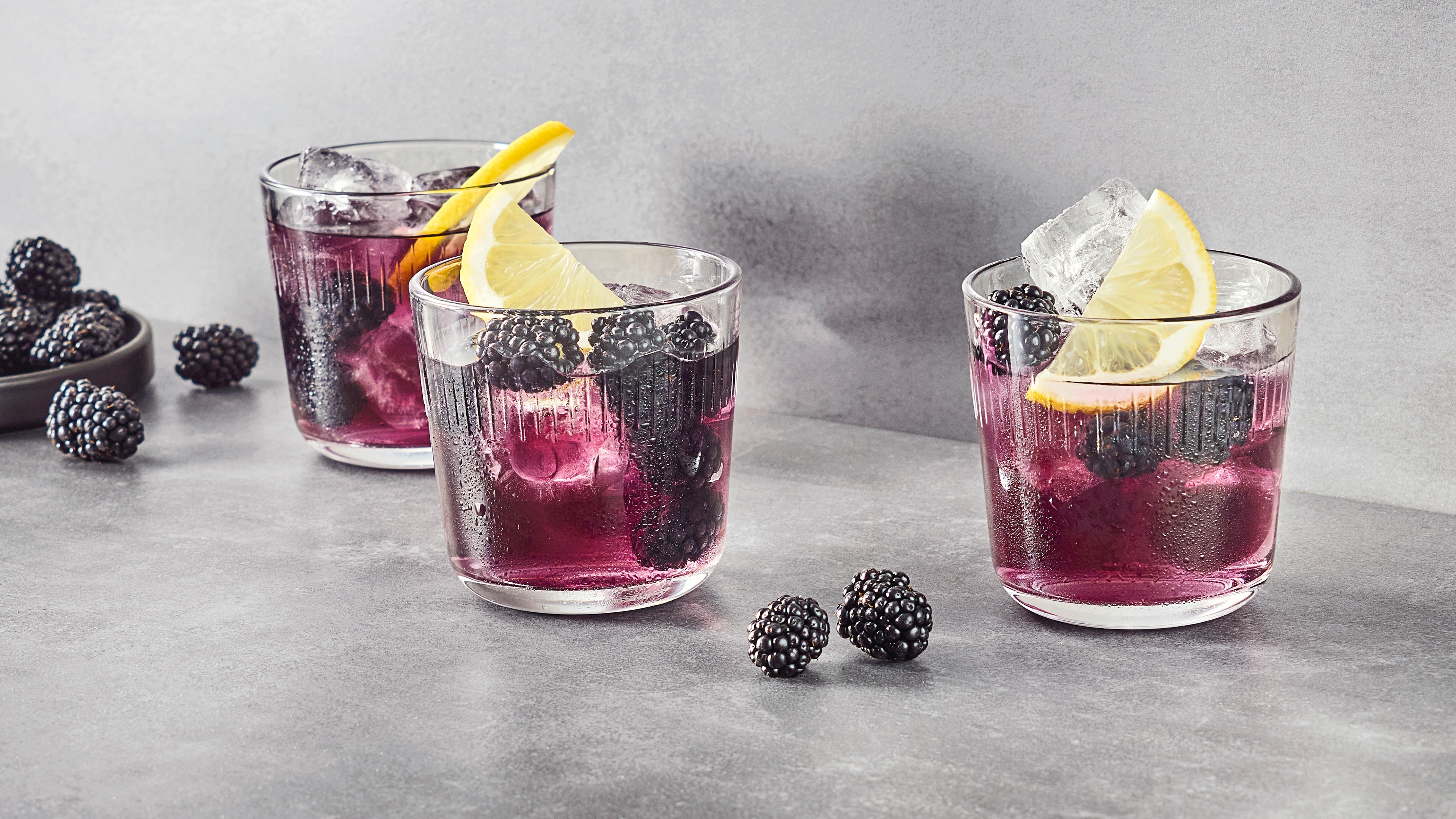 Drei durchsichtige Gläser mit violettem Eistee, gefüllt mit Brombeeren, Eiswürfeln und Zitronenschalen. Rundherum liegen weitere gefrorene Brombeeren.