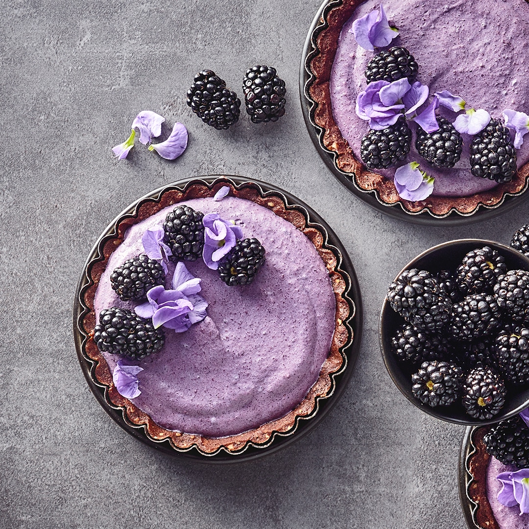Zwei Tartelettes mit Tortenboden und violetter Joghurtcreme, garniert mit Brombeeren und violetten Blüten. Daneben steht eine Schale mit weiteren Brombeeren.