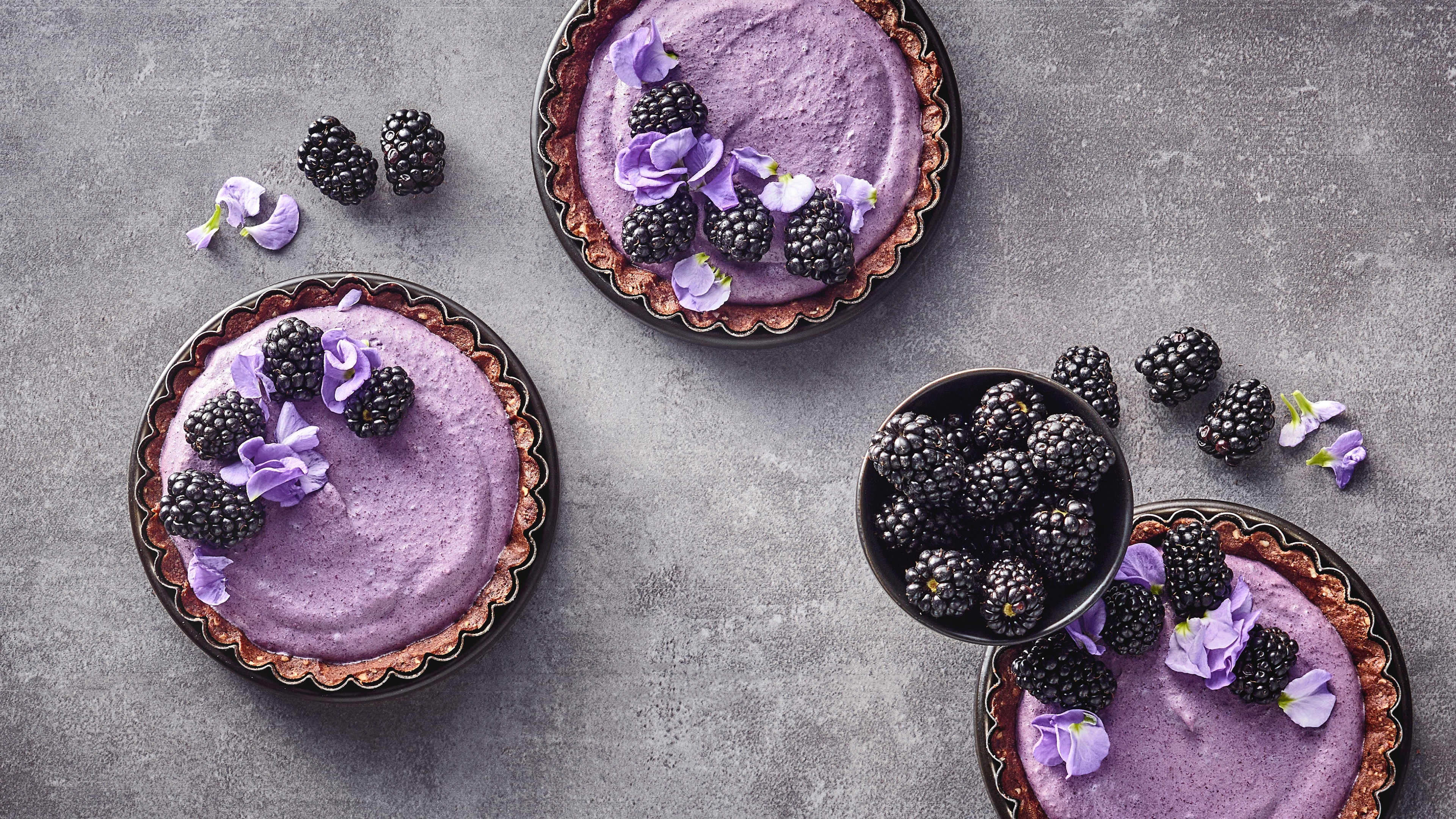 Drei Tartelettes mit Tortenboden und violetter Joghurtcreme, garniert mit Brombeeren und violetten Blüten. Dazwischen steht eine Schale mit weiteren Brombeeren.