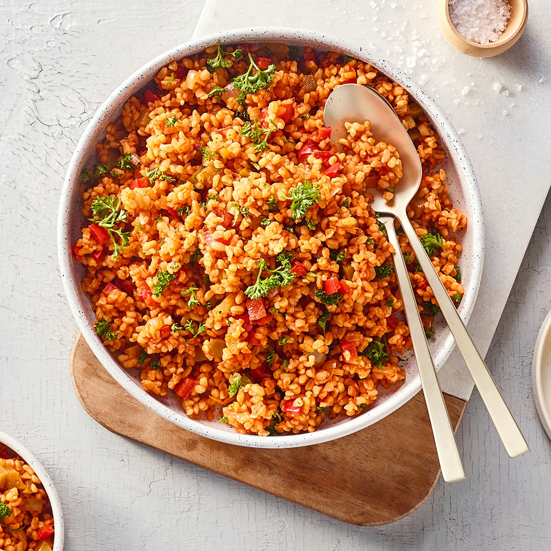 Eine Schüssel mit lockerem, orangefarbenem Bulgur, durchmischt mit kleinen roten Stücken und grünen Kräutern.