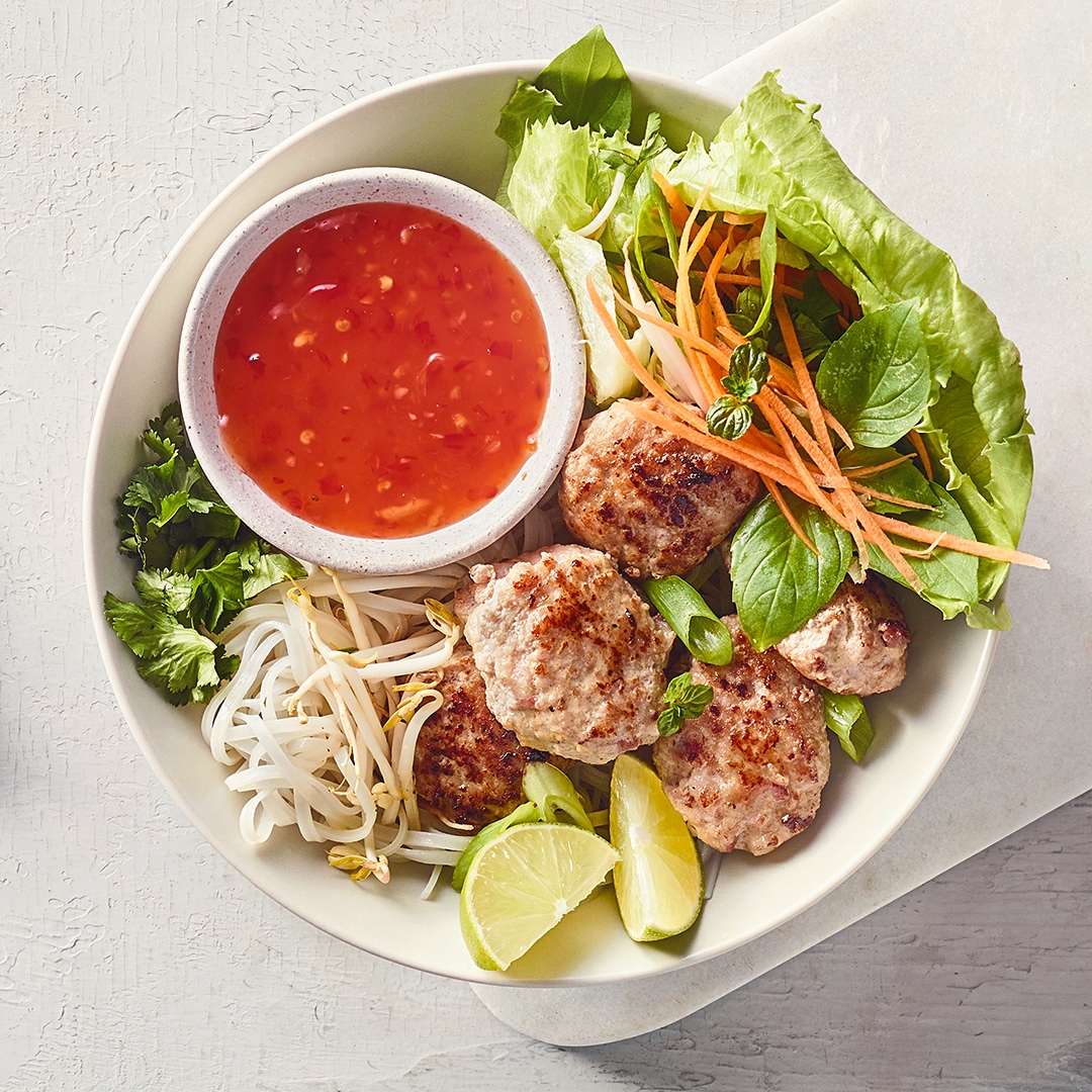 Eine Schale mit gebratenen Fleischbällchen, frischen Salatblättern, Karottenstreifen, Limettenstücken und roter Sauce