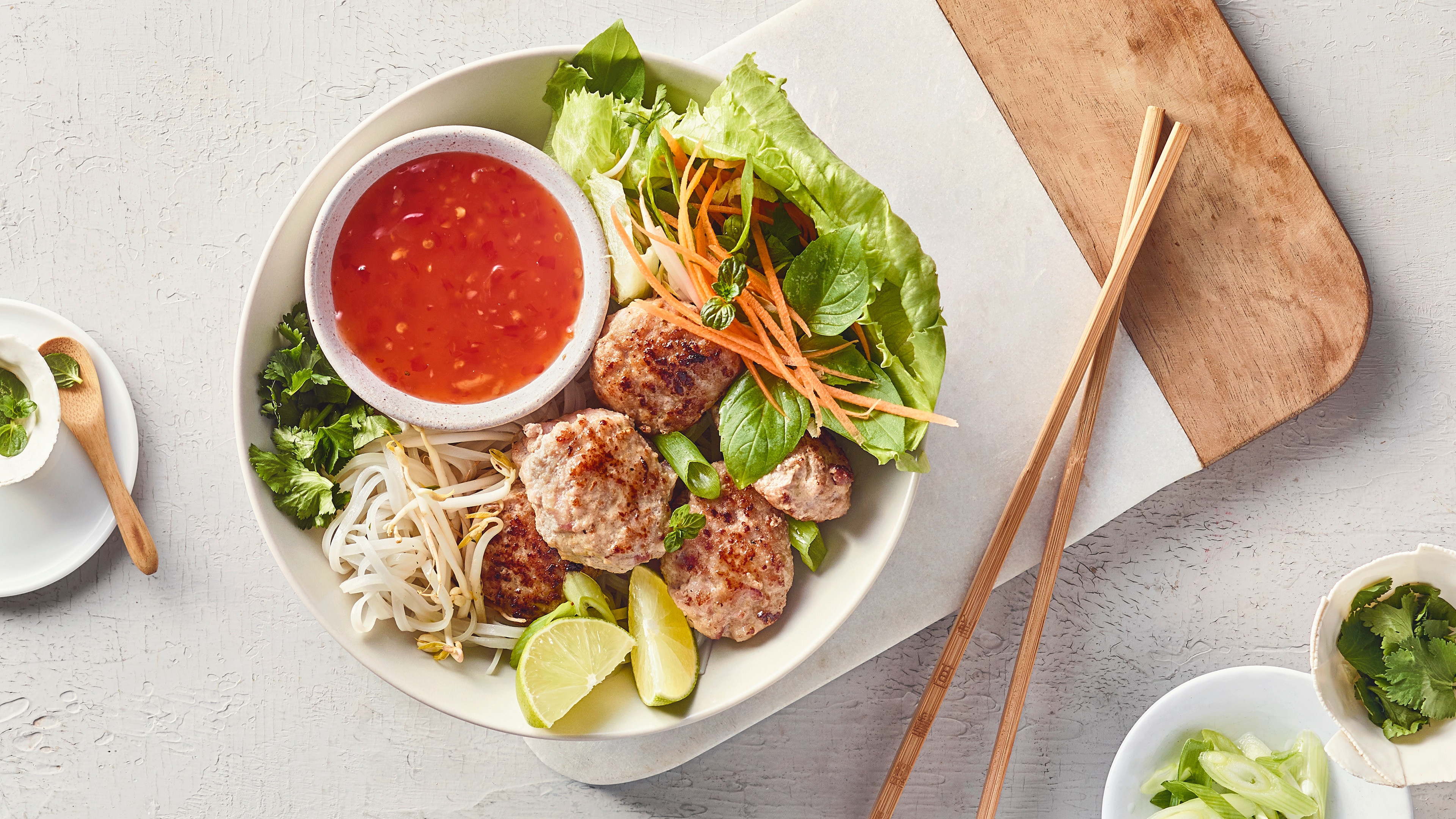 Eine Schale mit gebratenen Fleischbällchen, frischen Salatblättern, Karottenstreifen, Limettenstücken und roter Sauce