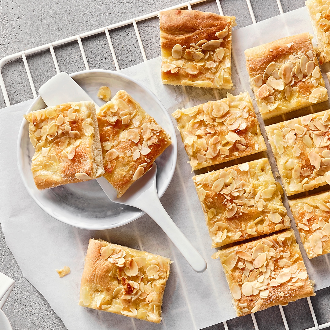 Ein Teller mit mehreren rechteckigen Kuchenstücken, belegt mit hellen Mandelblättchen auf einem goldenen Teig.