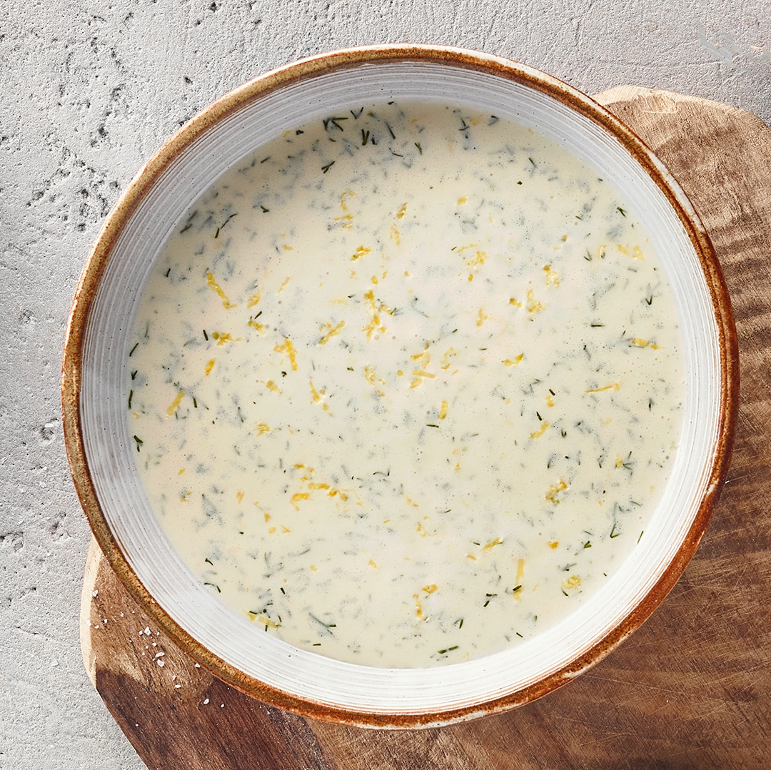 Eine Schale mit heller, cremiger Flüssigkeit, durchzogen von feinen grünen Kräuterstücken und kleinen gelben Streifen.
