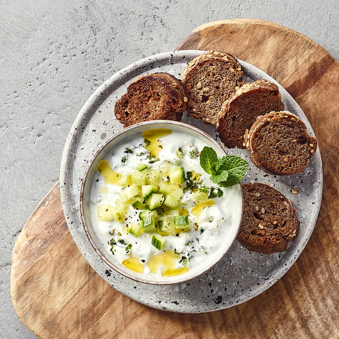 Ein Teller mit weißem Joghurt, grünen Gurkenwürfeln, grünem Öl und Kräutern, dazu braune Brotscheiben.