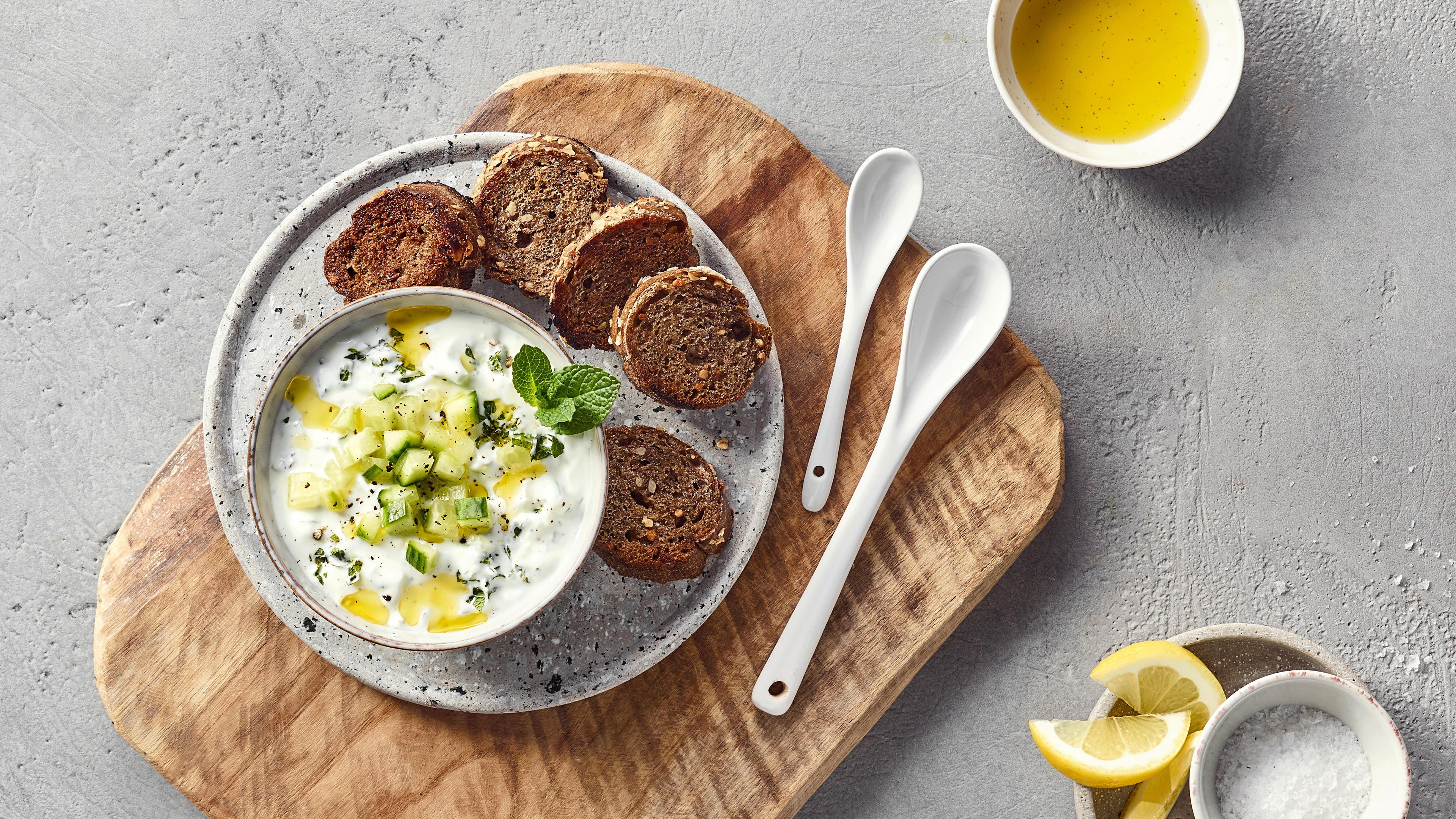 Ein Teller mit weißem Joghurt, grünen Gurkenwürfeln, grünem Öl und Kräutern, dazu braune Brotscheiben.