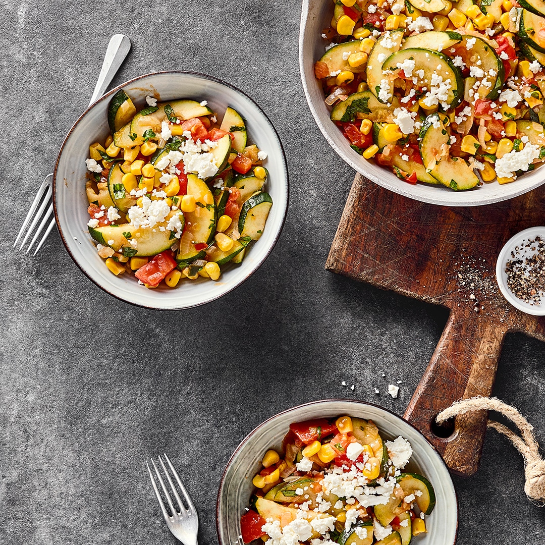 Runde Schälchen mit Mais, Zucchini- und Paprikastückchen, getoppt mit Schafskäsebröseln und gehacktem Koriander
