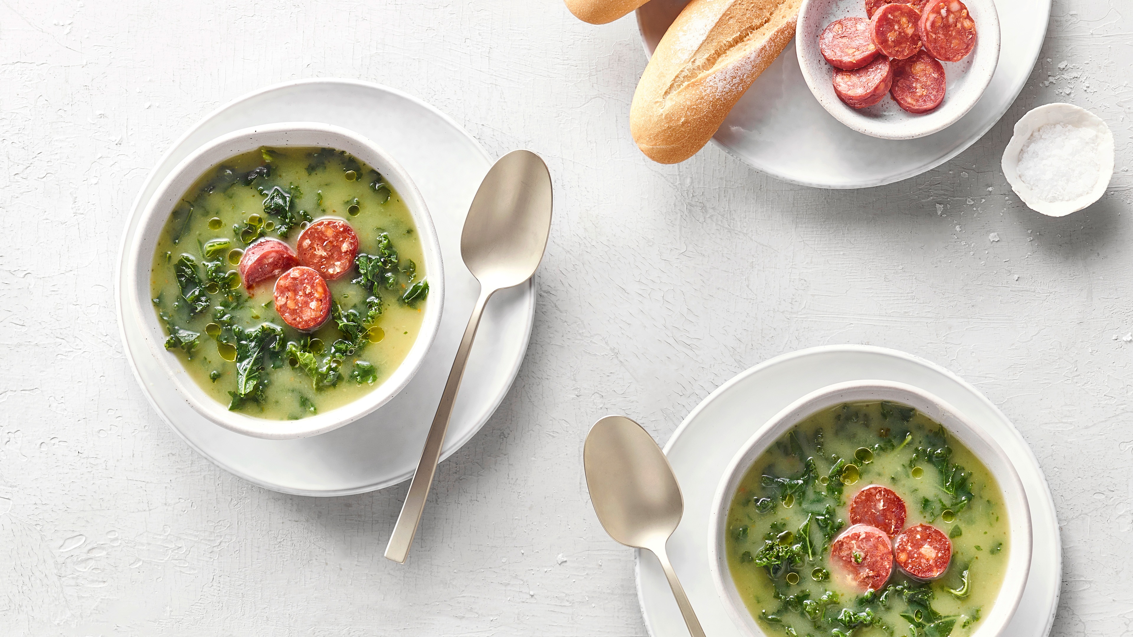 Zwei kleine Schüsseln mit grüner, kohliger Suppe und kleinen, runden Fleischscheiben. Oben rechts befinden sich Baguettebrot, weitere kleine Fleischscheiben und Salz.