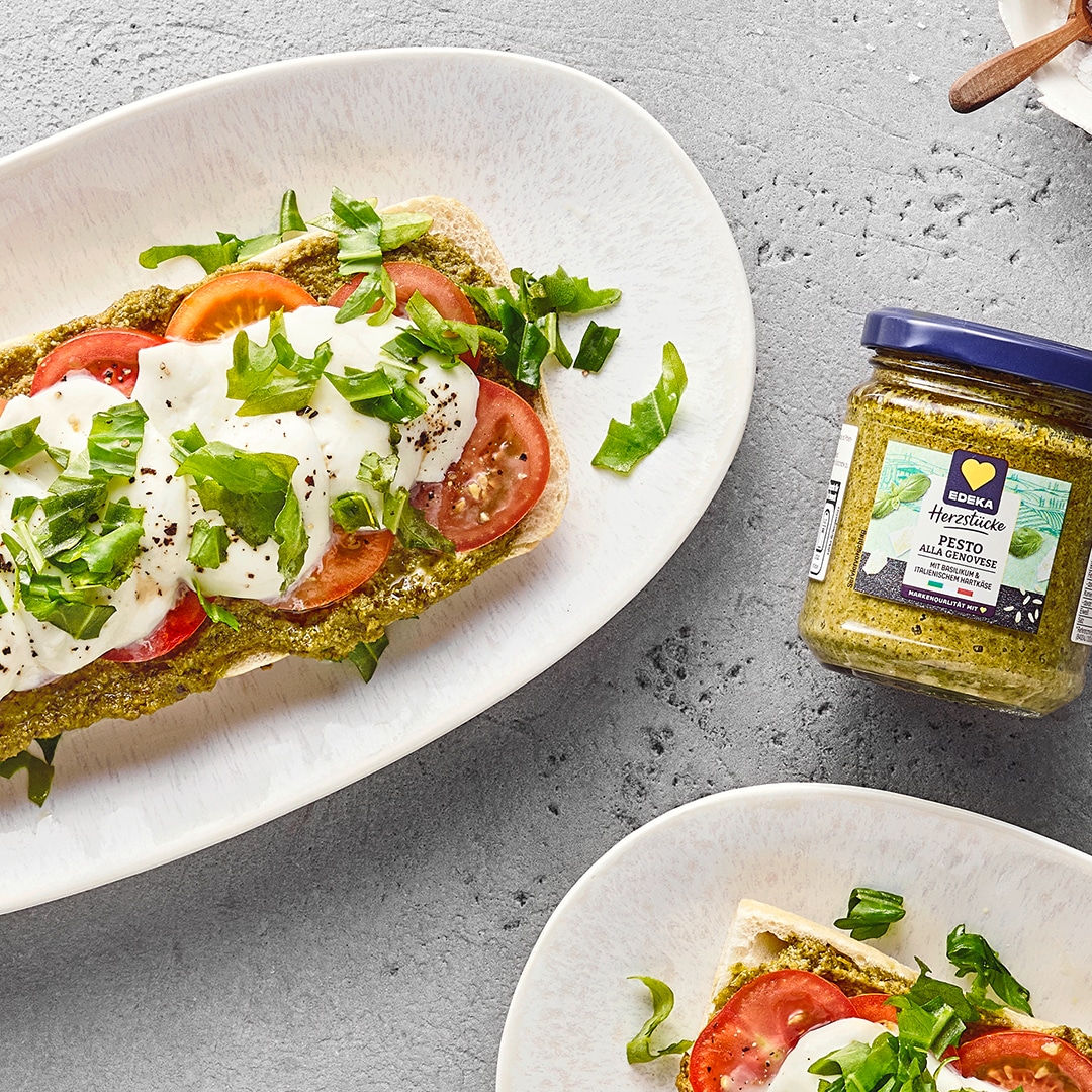 Geröstetes Ofensandwich mit geschmolzenem Mozzarella, Tomatenscheiben, Pesto und Basilikum. Daneben ein Glas EDEKA Herzstücke Pesto alla Genovese.
