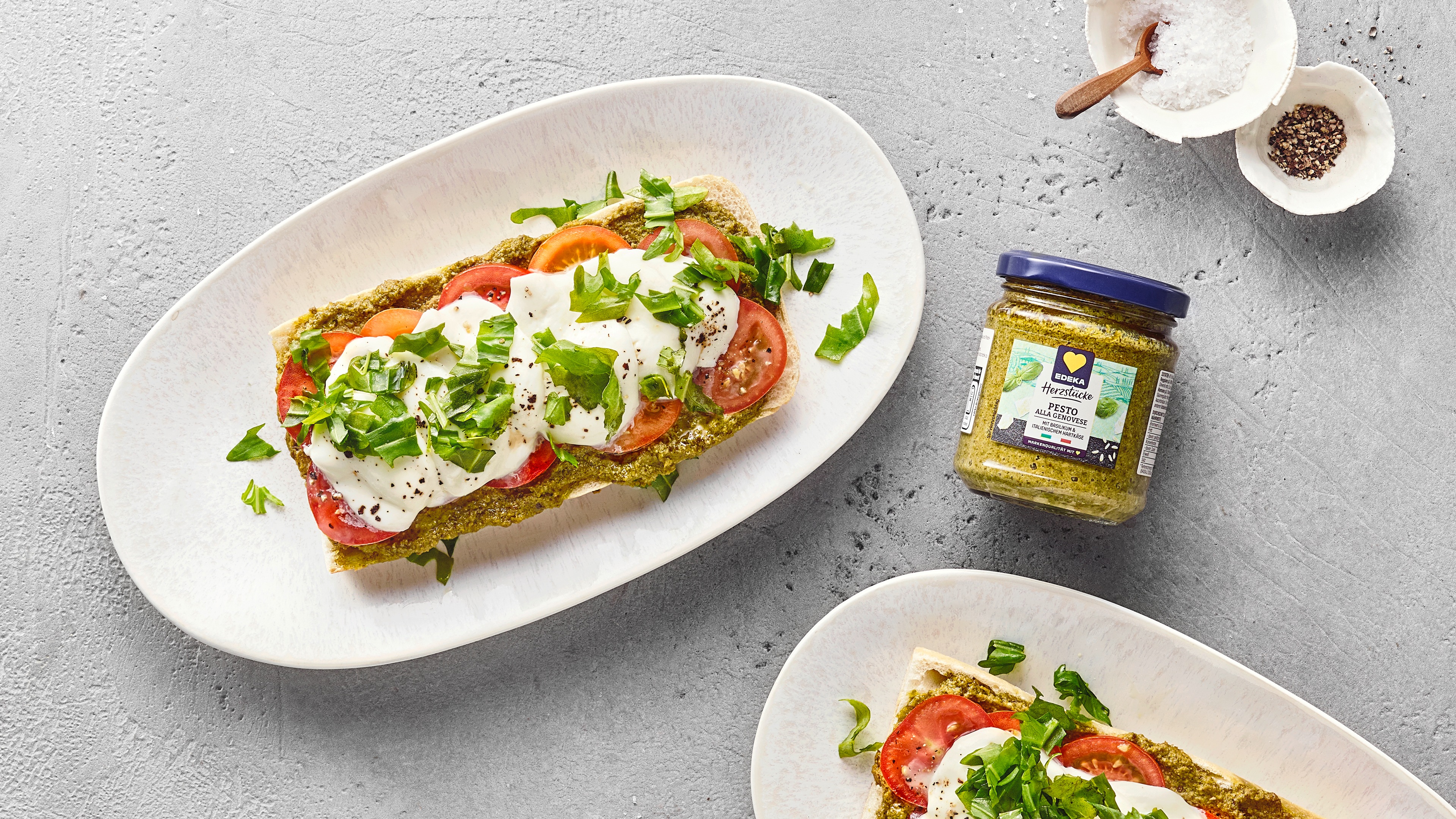 Geröstete Ofensandwiches mit geschmolzenem Mozzarella, Tomatenscheiben, Pesto und Basilikum. Daneben ein Glas EDEKA Herzstücke Pesto alla Genovese.