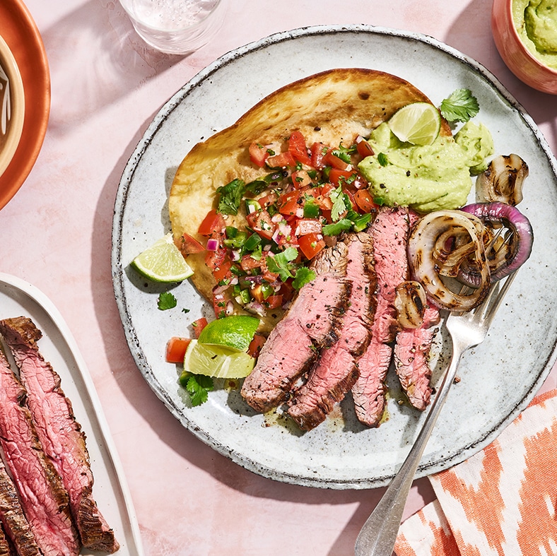 Runder Teller mit einer Tortilla, belegt mit gewürfelten Tomaten, Zwiebeln, Petersilie, Limettenstücken, in Scheiben geschnittenem Rindfleisch und Guacamole, dazu eine Gabel. Links unten steht ein weiterer Teller, auf dem zusätzlich in Scheiben geschnittenes Rindfleisch liegt.