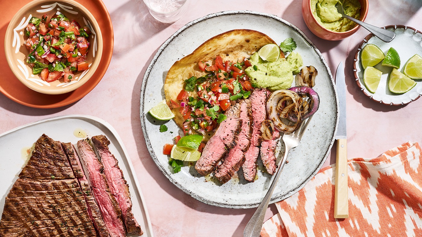 Runder Teller mit einer Tortilla, belegt mit gewürfelten Tomaten, Zwiebeln, Petersilie, Limettenstücken, in Scheiben geschnittenem Rindfleisch und Guacamole, dazu eine Gabel und ein Messer. Links oben steht eine runde Schale mit Tomatensalat. Rechts oben steht eine Schale mit Guacamole und eine Schale mit Limettenstücken. Links unten steht ein weiterer Teller, auf dem zusätzlich in Scheiben geschnittenes Rindfleisch liegt. Rechts unten liegt ein Küchentuch.