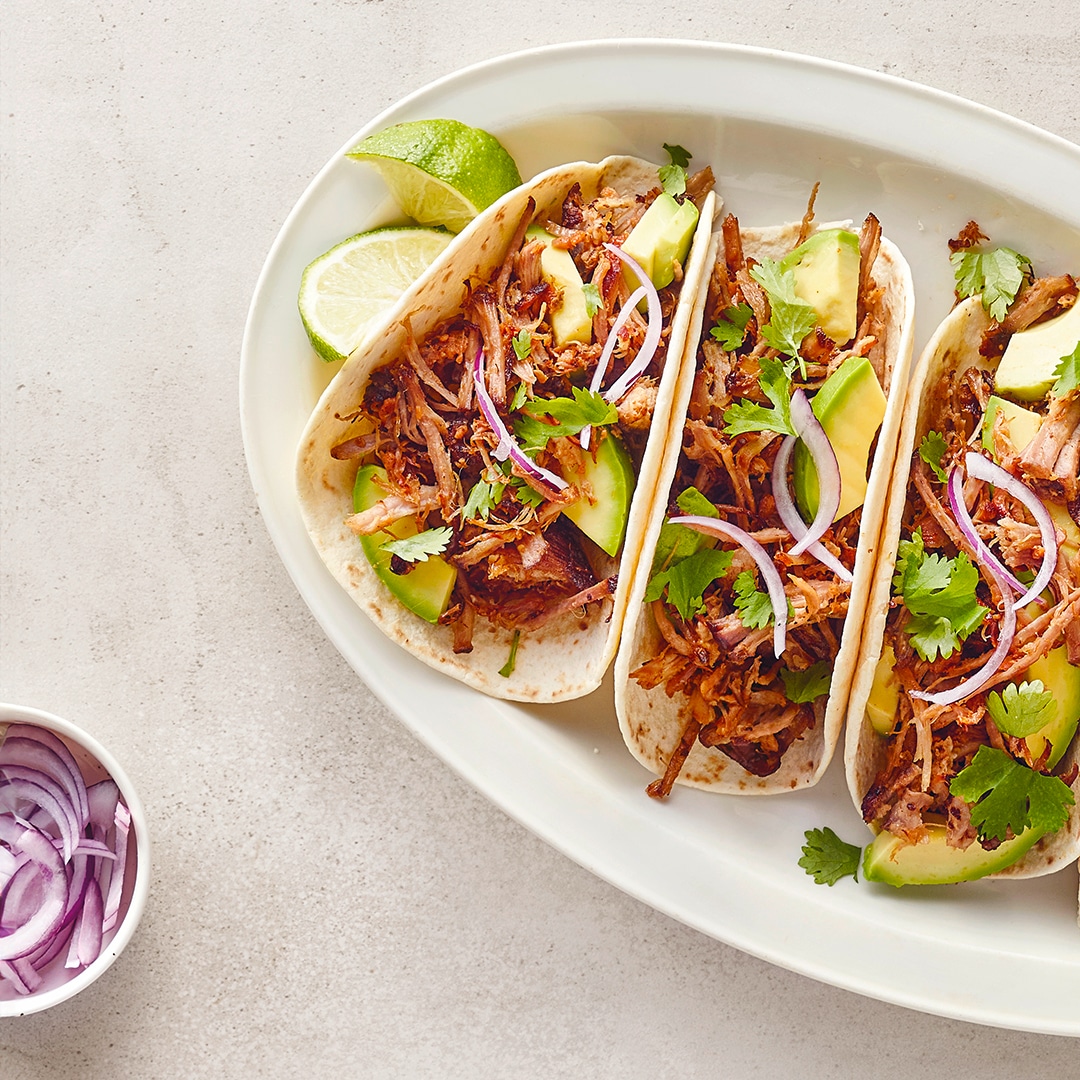 Ein Teller mit vier Tacos, gefüllt mit Fleisch, Avocado, roten Zwiebeln und Koriander, dazu Limettenstücke