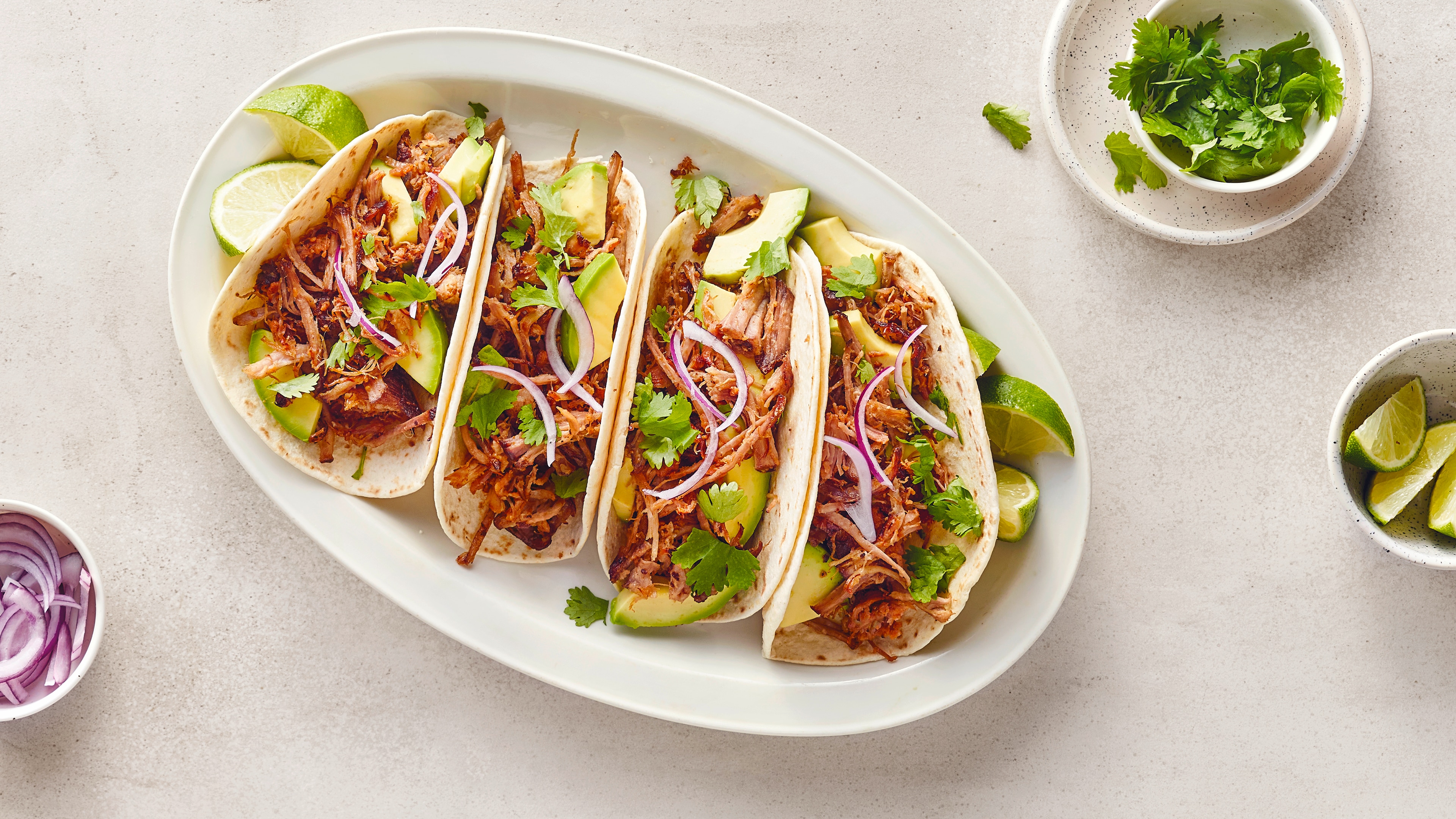 Rezept für Carnitas Ein Teller mit vier Tacos, gefüllt mit Fleisch, Avocado, roten Zwiebeln und Koriander, dazu Limettenstücke