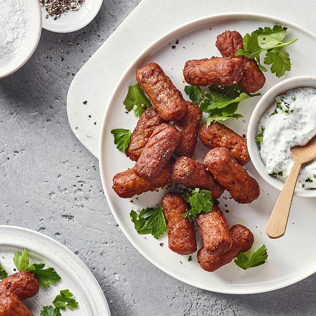 Ein Teller mit gebratenen braunen Würstchen, garniert mit grünen Kräutern und einer Schale weißer Creme