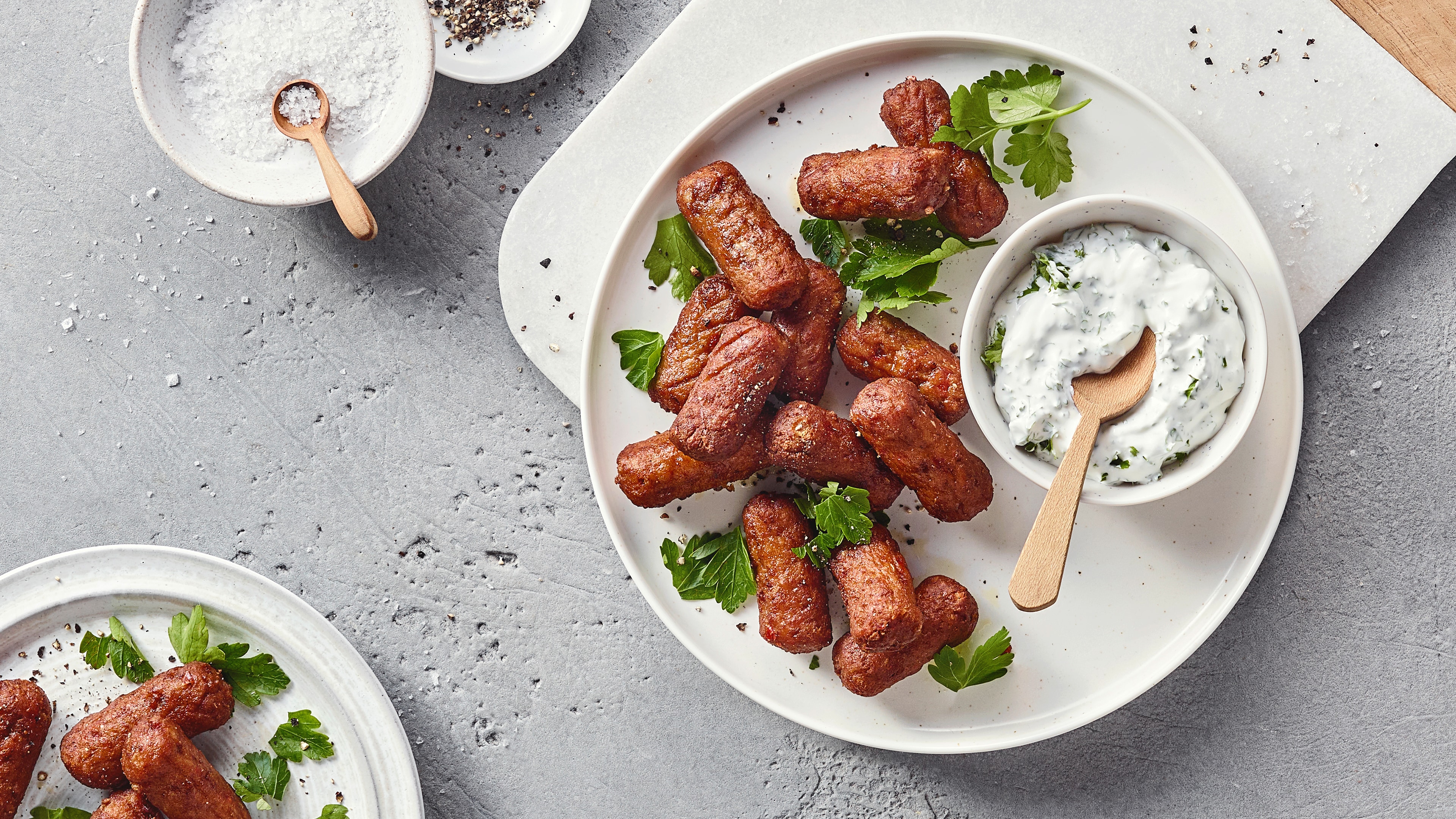 Ein Teller mit gebratenen braunen Würstchen, garniert mit grünen Kräutern und einer Schale weißer Creme