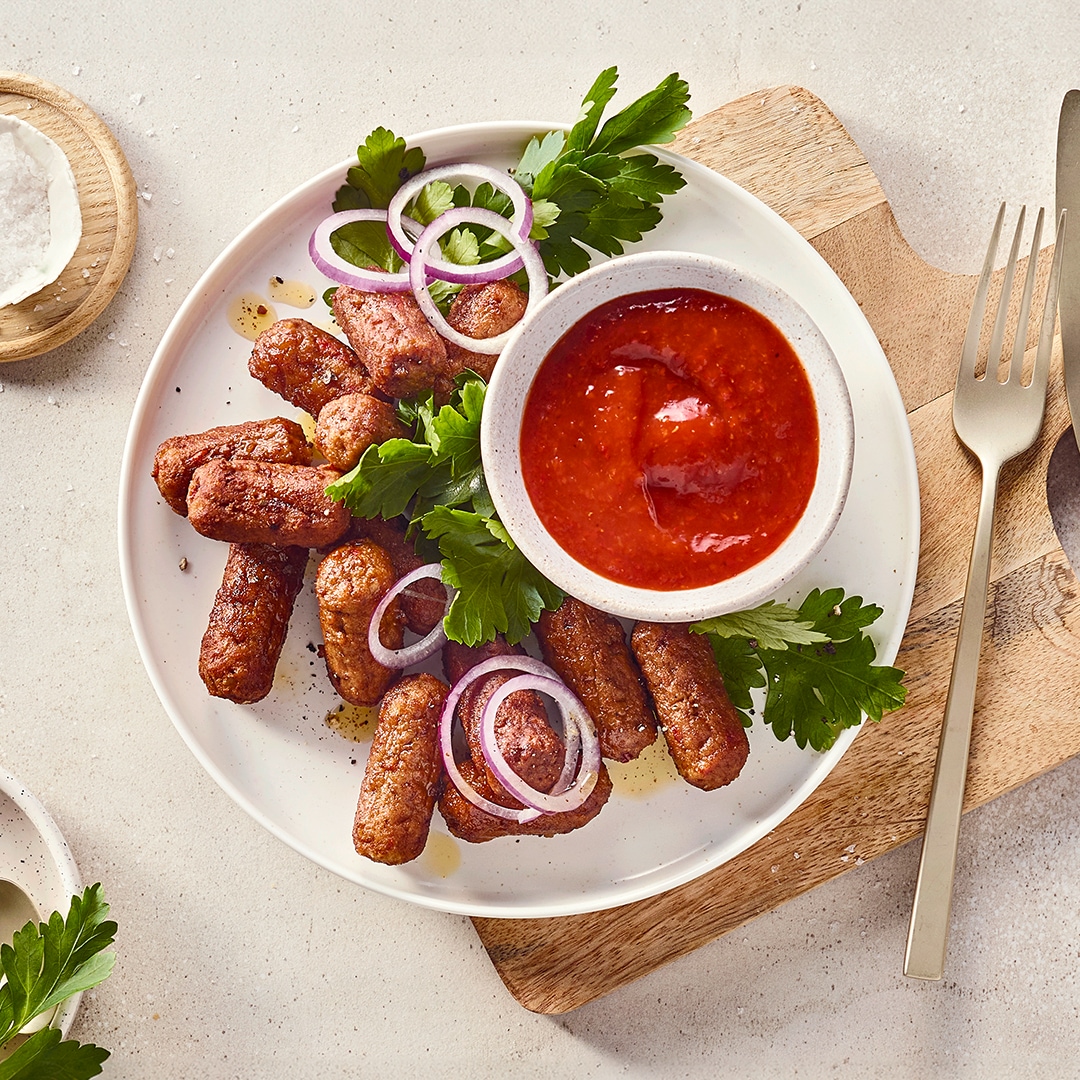 Ein Teller mit gebratenen Cevapcici, garniert mit Petersilie und roten Zwiebelringen, daneben eine Schale mit roter Soße.