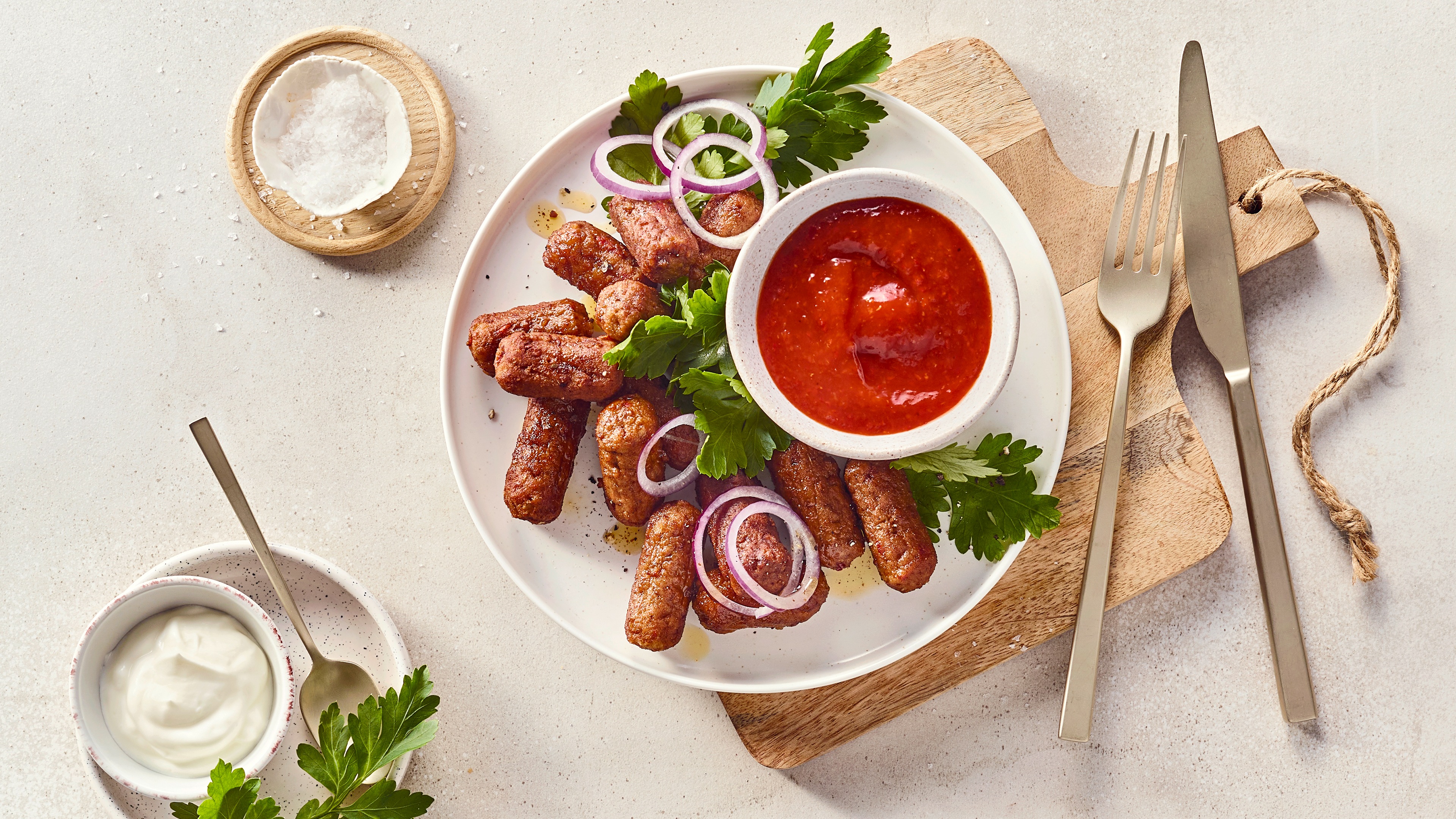 Ein Teller mit gebratenen Cevapcici, garniert mit Petersilie und roten Zwiebelringen, daneben eine Schale mit roter Soße.