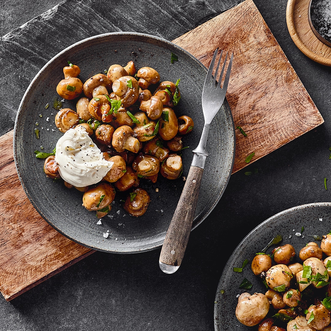 Ein rustikaler Teller mit kleinen Champignons, getoppt mit einem Klecks weißer creme und frischen gehackten Kräutern