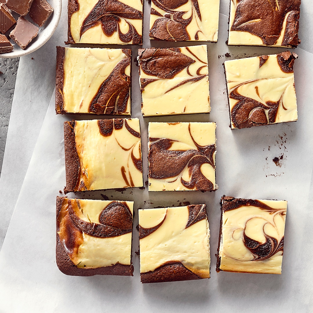 Viereckig geschnittene Brownies aus hellem und dunklem Teig auf Backpapier, daneben eine Schale mit Schokolade