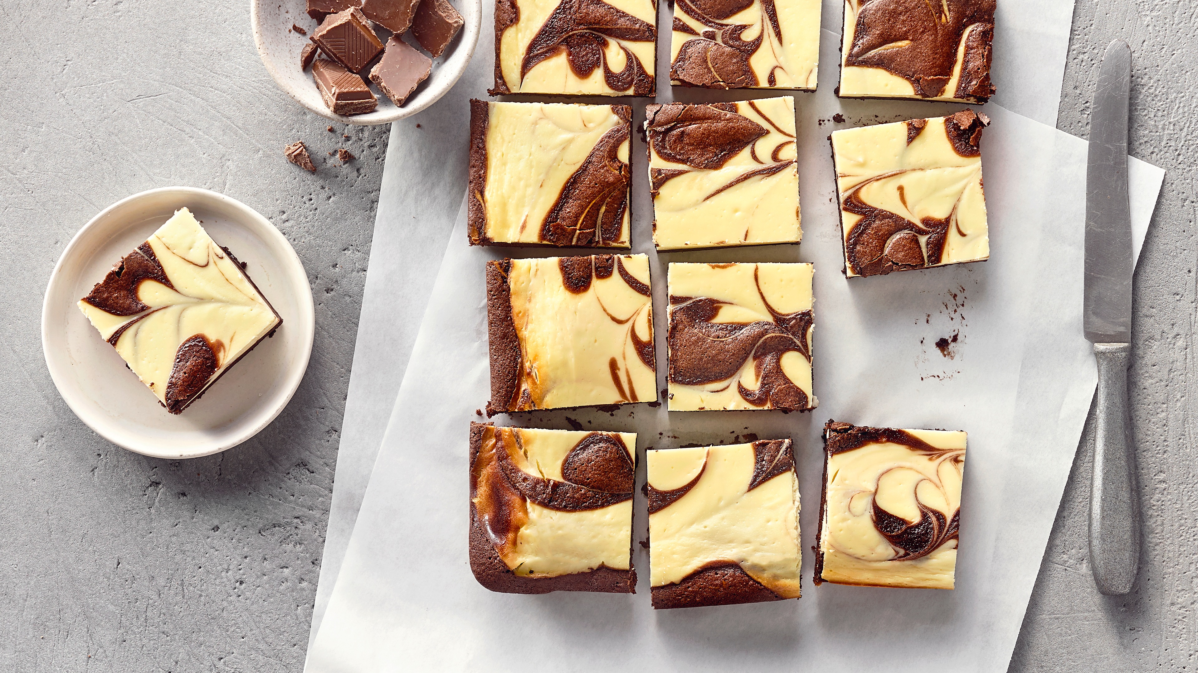 Viereckig geschnittene Brownies aus hellem und dunklem Teig auf Backpapier, daneben eine Schale mit Schokolade