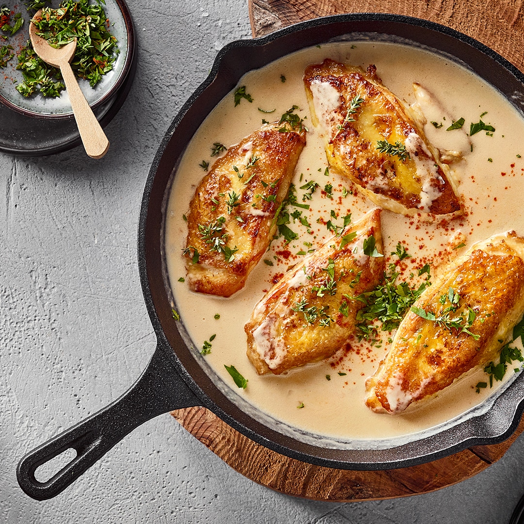 Eine Pfanne mit vier gebratenen Hähnchenbrustfilets in heller Soße, bestreut mit grünen Kräutern.
