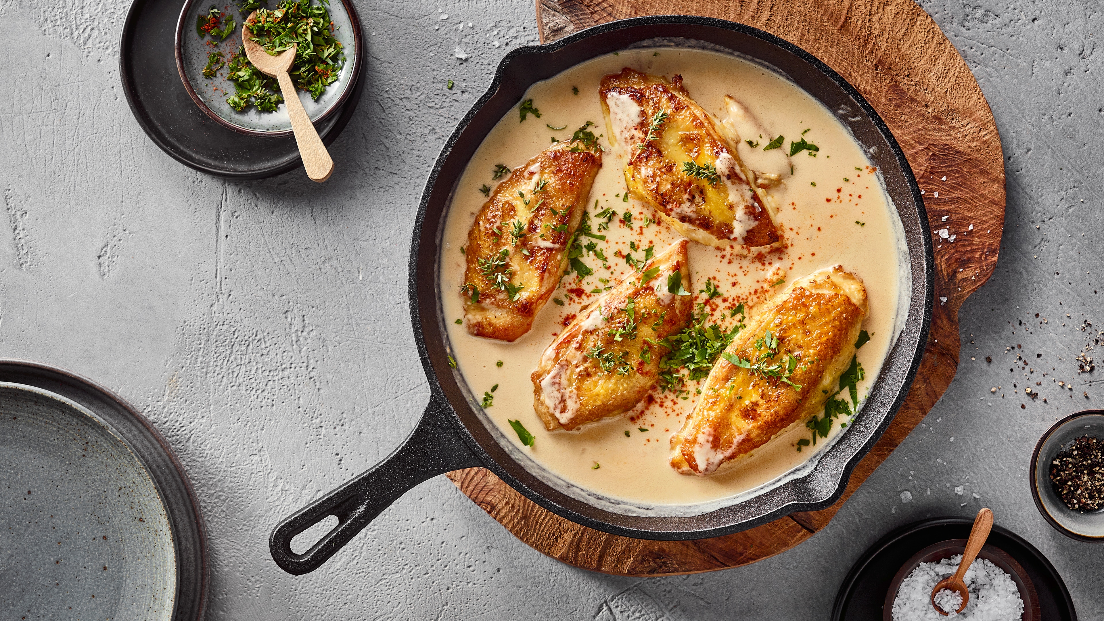Eine Pfanne mit vier gebratenen Hähnchenbrustfilets in heller Soße, bestreut mit grünen Kräutern.