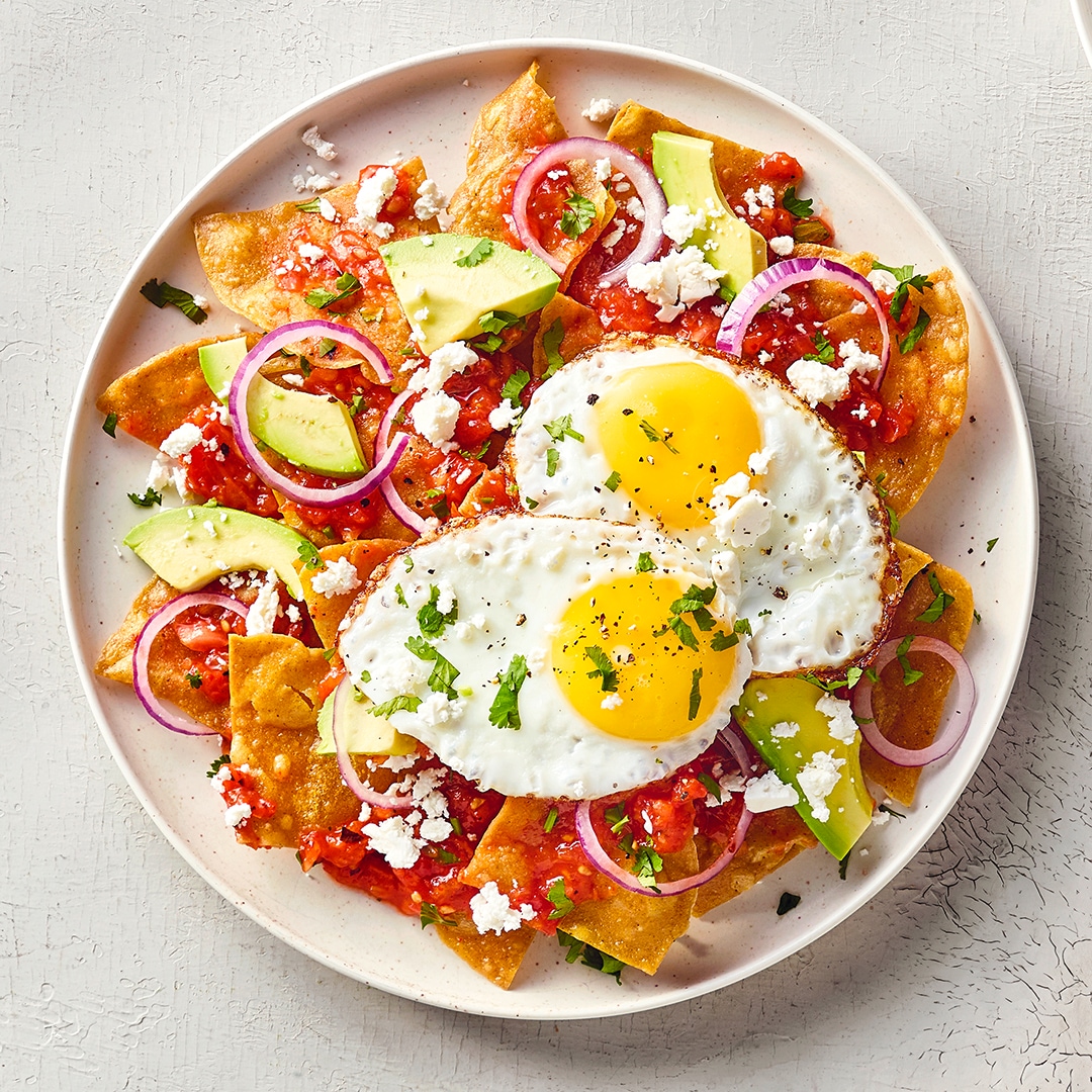 Ein Teller mit Tortillastücken, roter Sauce, zwei Spiegeleiern, Avocadostücken, roten Zwiebelringen und zerbröseltem Käse