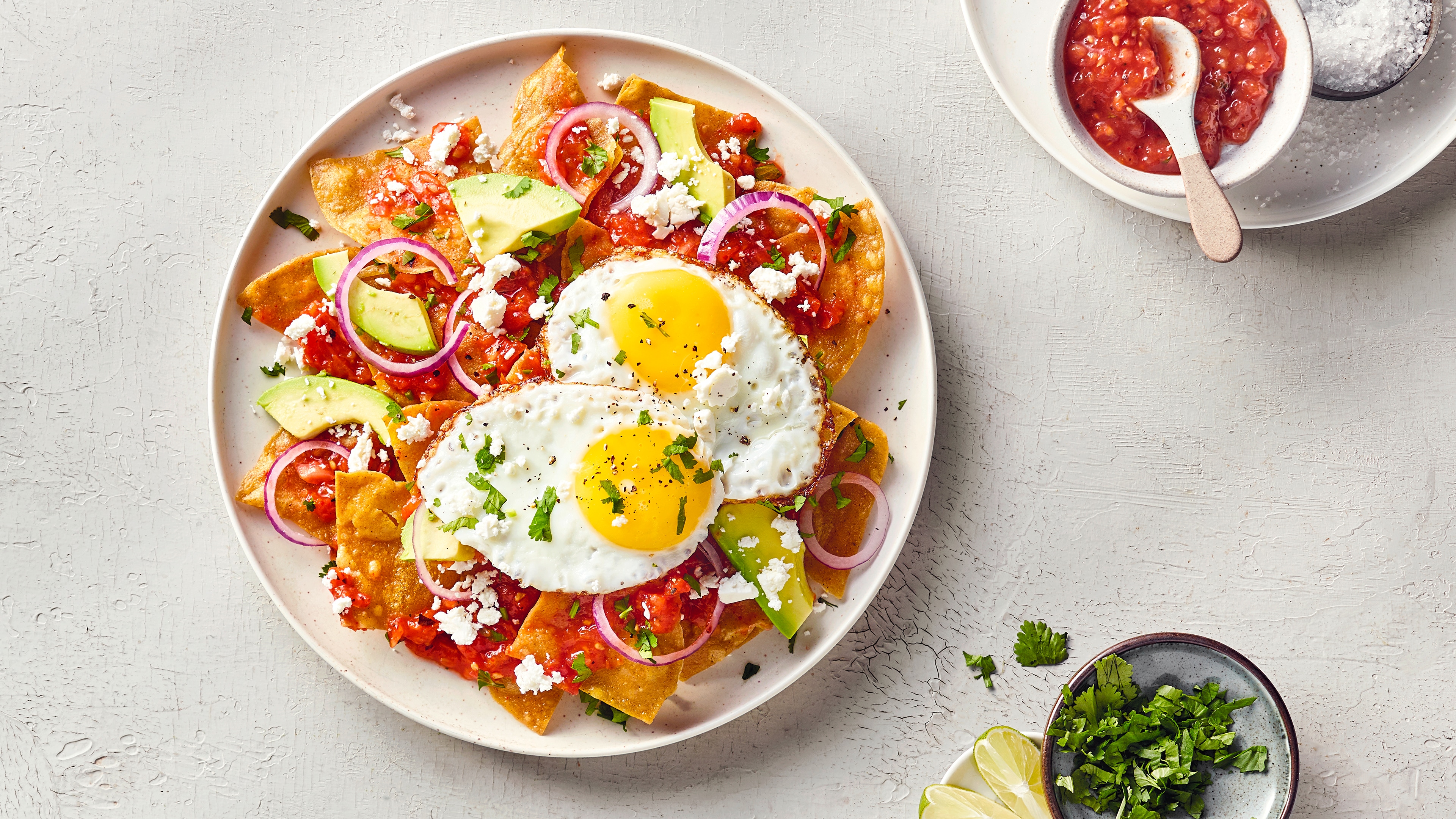 Ein Teller mit Tortillastücken, roter Sauce, zwei Spiegeleiern, Avocadostücken, roten Zwiebelringen und zerbröseltem Käse