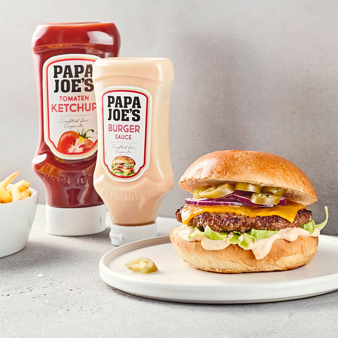 Burger mit Fleisch, Käse, Salat, Jalapenos und Soße, daneben Papa Joes Burger-Sauce und Ketchup