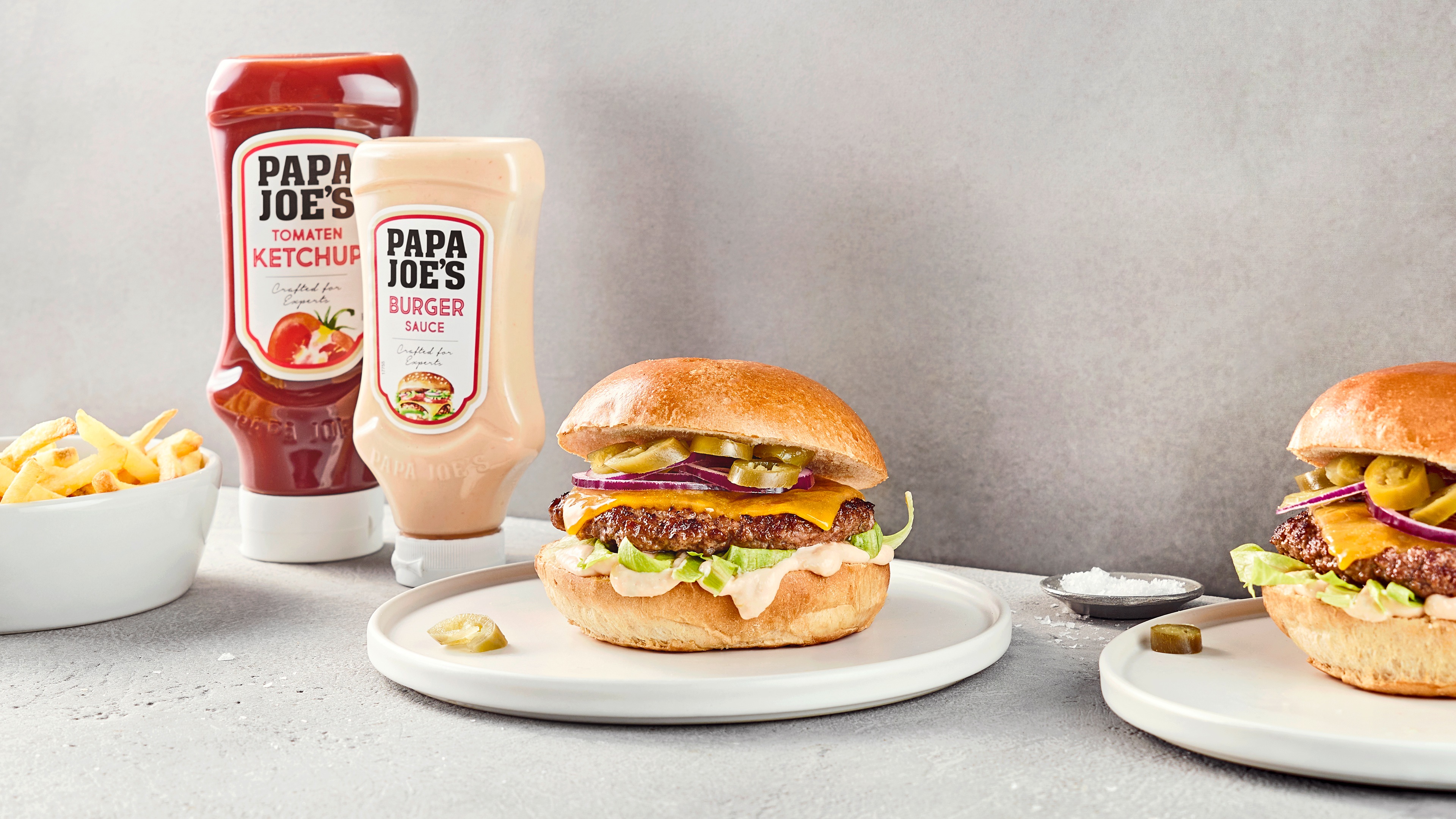 Zwei Burger mit Fleisch, Käse, Salat, Jalapenos und Soße, daneben Papa Joes Burger-Sauce und Ketchup