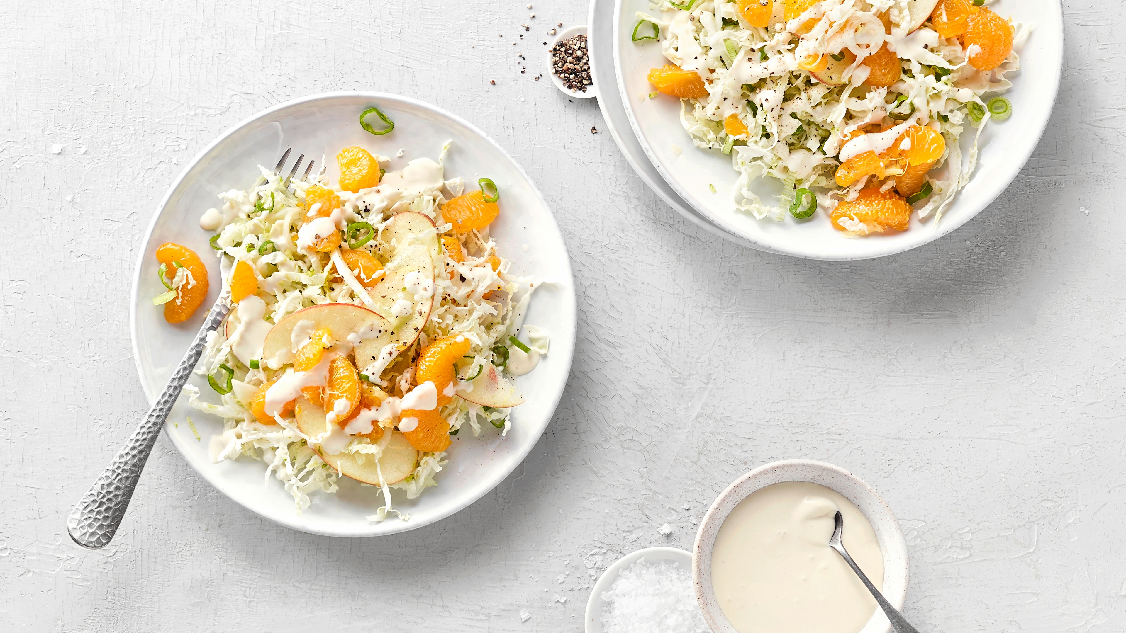 Zwei Teller mit Kohlsalat, Mandarinen, Apfelstücken und Joghurtdressing. Unten rechts eine Schale mit mehr Joghurtdressing.