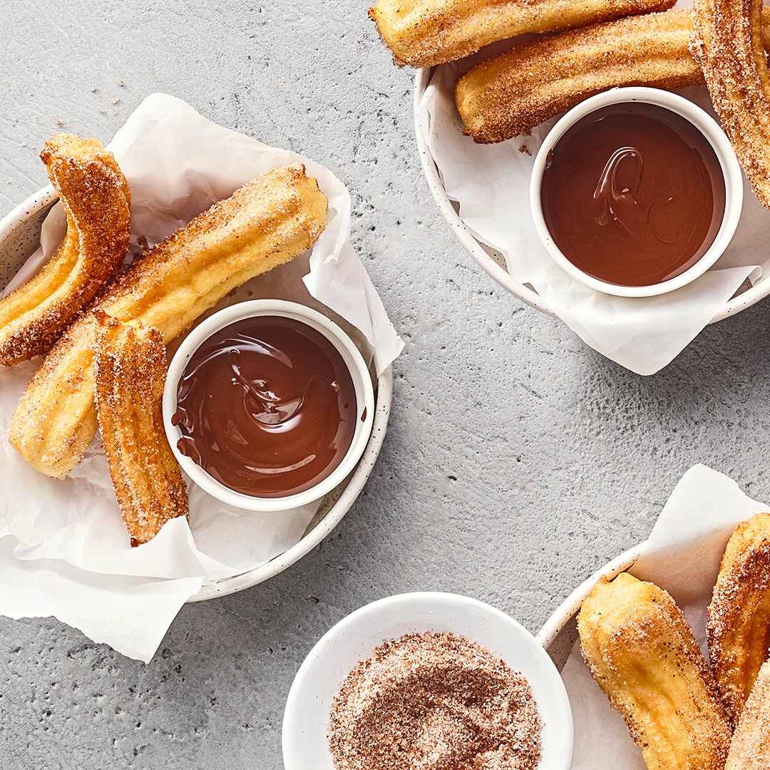 Drei kleine Schüsseln mit zuckrigen Churros auf Papiertüchern. Dazu zwei Dippschalen mit flüssiger Schokolade und eine weitere mit Zimt und Zucker.