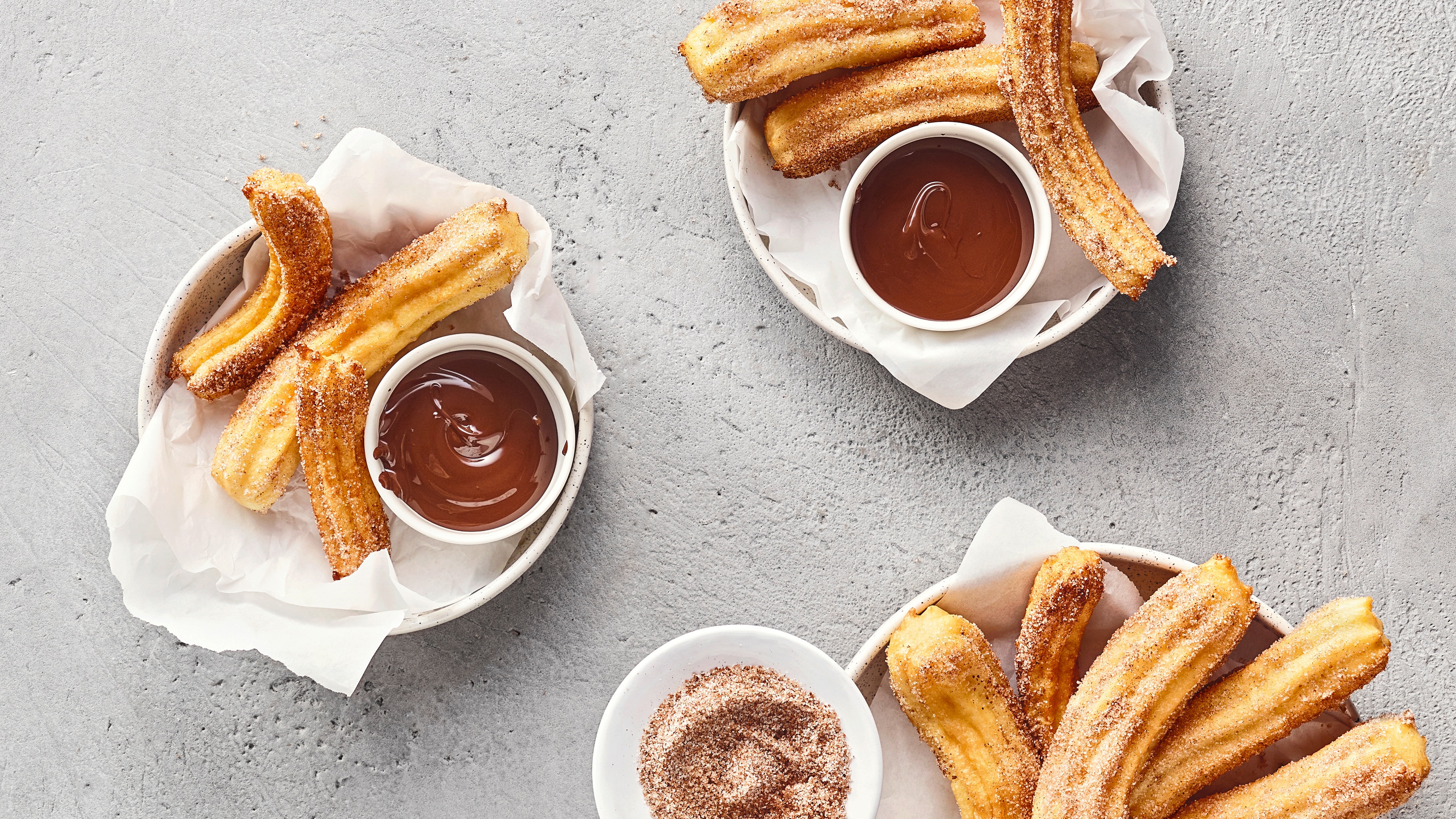 Drei kleine Schüsseln mit zuckrigen Churros auf Papiertüchern. Dazu zwei Dippschalen mit flüssiger Schokolade und eine weitere mit Zimt und Zucker.