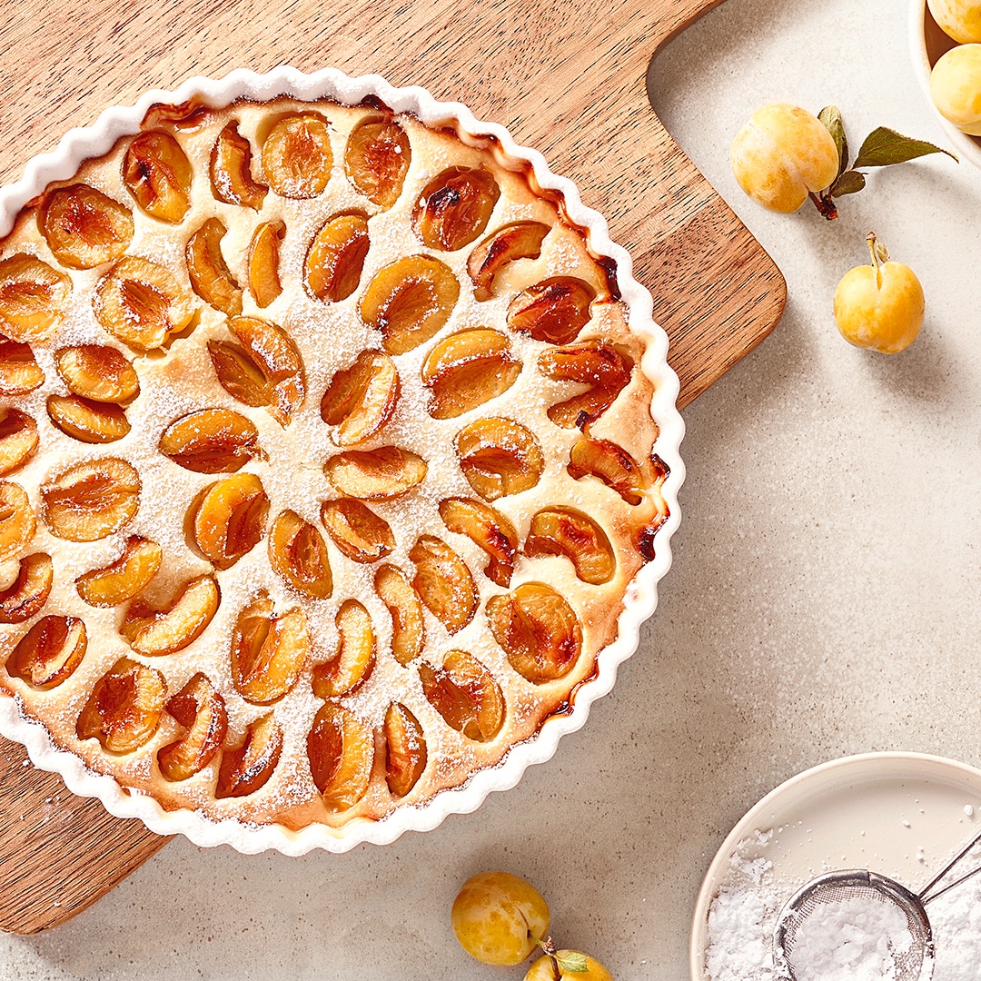 Mirabellenkuchen in einer runden Tarteform auf einem Holzbrett, mit frischen Mirabellen und Puderzucker daneben.