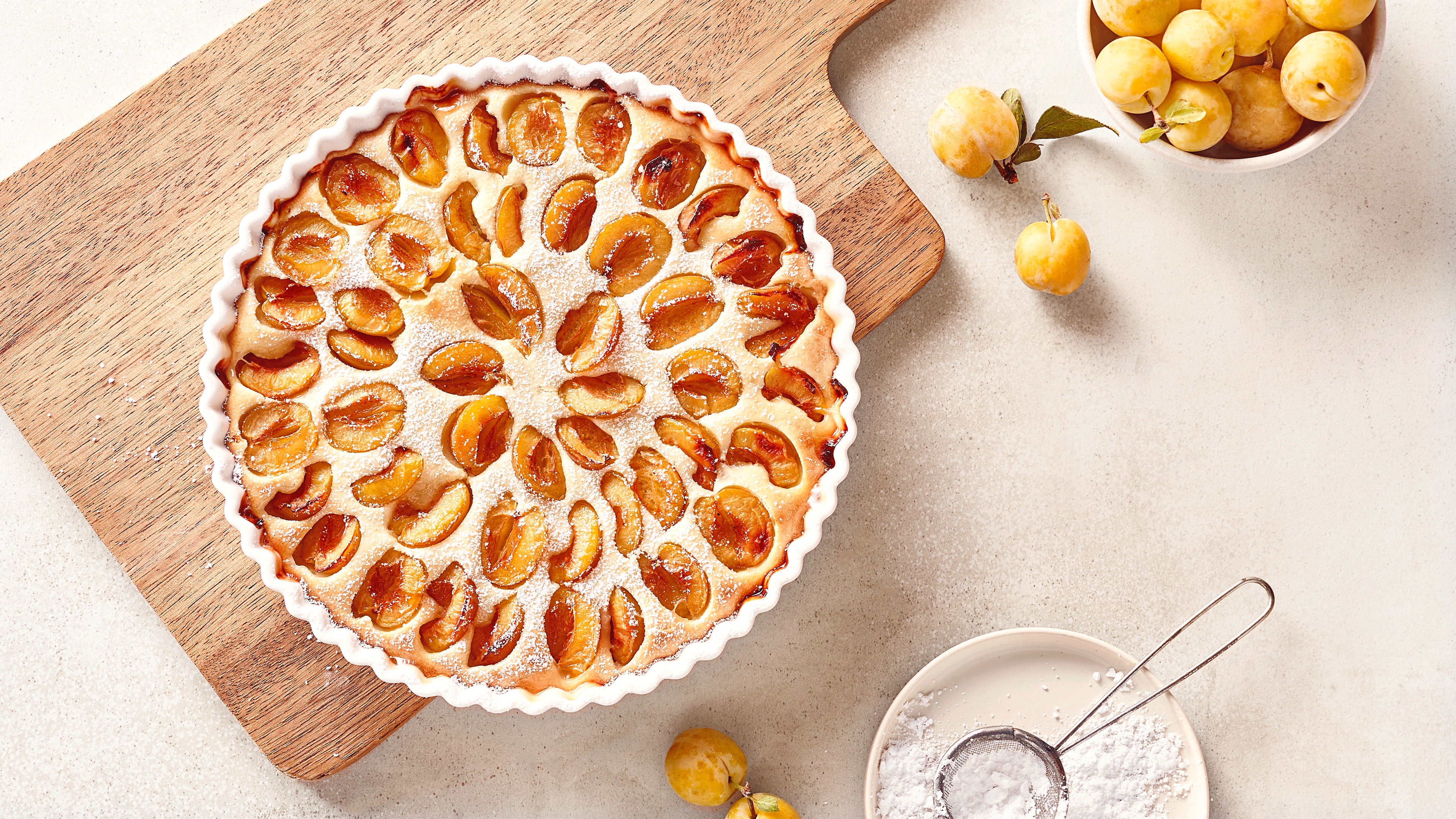 Mirabellenkuchen in einer runden Tarteform auf einem Holzbrett, mit frischen Mirabellen und Puderzucker daneben.