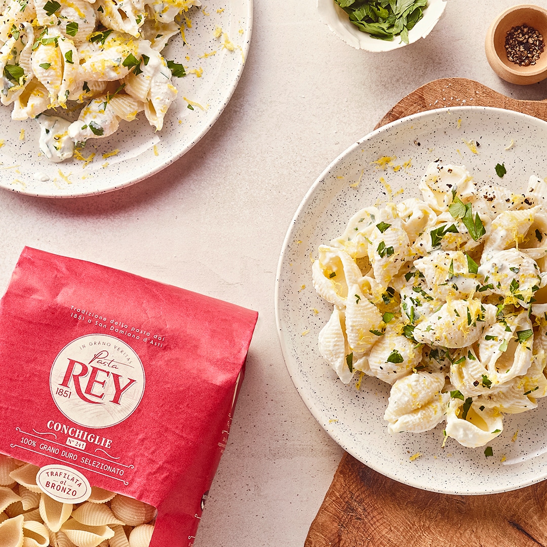 Cremige Muschelnudeln mit Zitronenabrieb und frischen Kräutern auf hellem Teller; daneben Pasta Rey Conchiglie Verpackung.