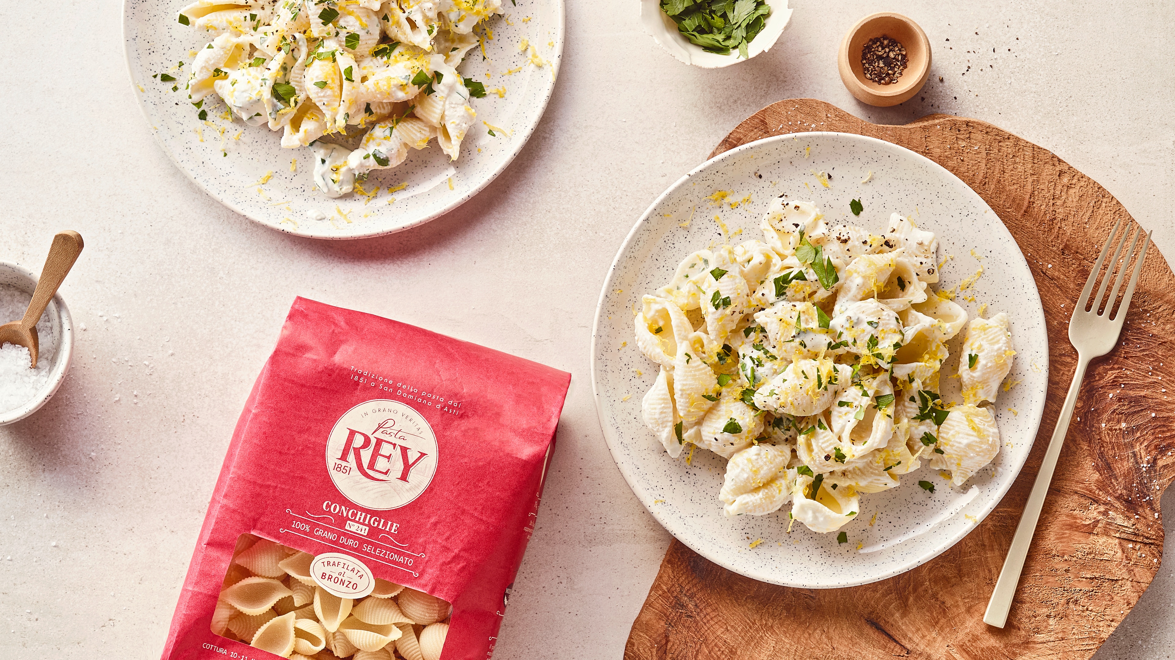 Cremige Muschelnudeln mit Zitronenabrieb und frischen Kräutern auf hellen Tellern; daneben Pasta Rey Conchiglie Verpackung.
