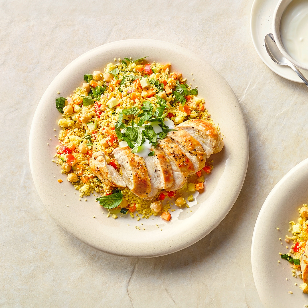 Ein Teller mit Couscous, Kichererbsen, Gurke, Paprika und Karotte, getoppt mit gebratenem Hähnchen und weißer Soße