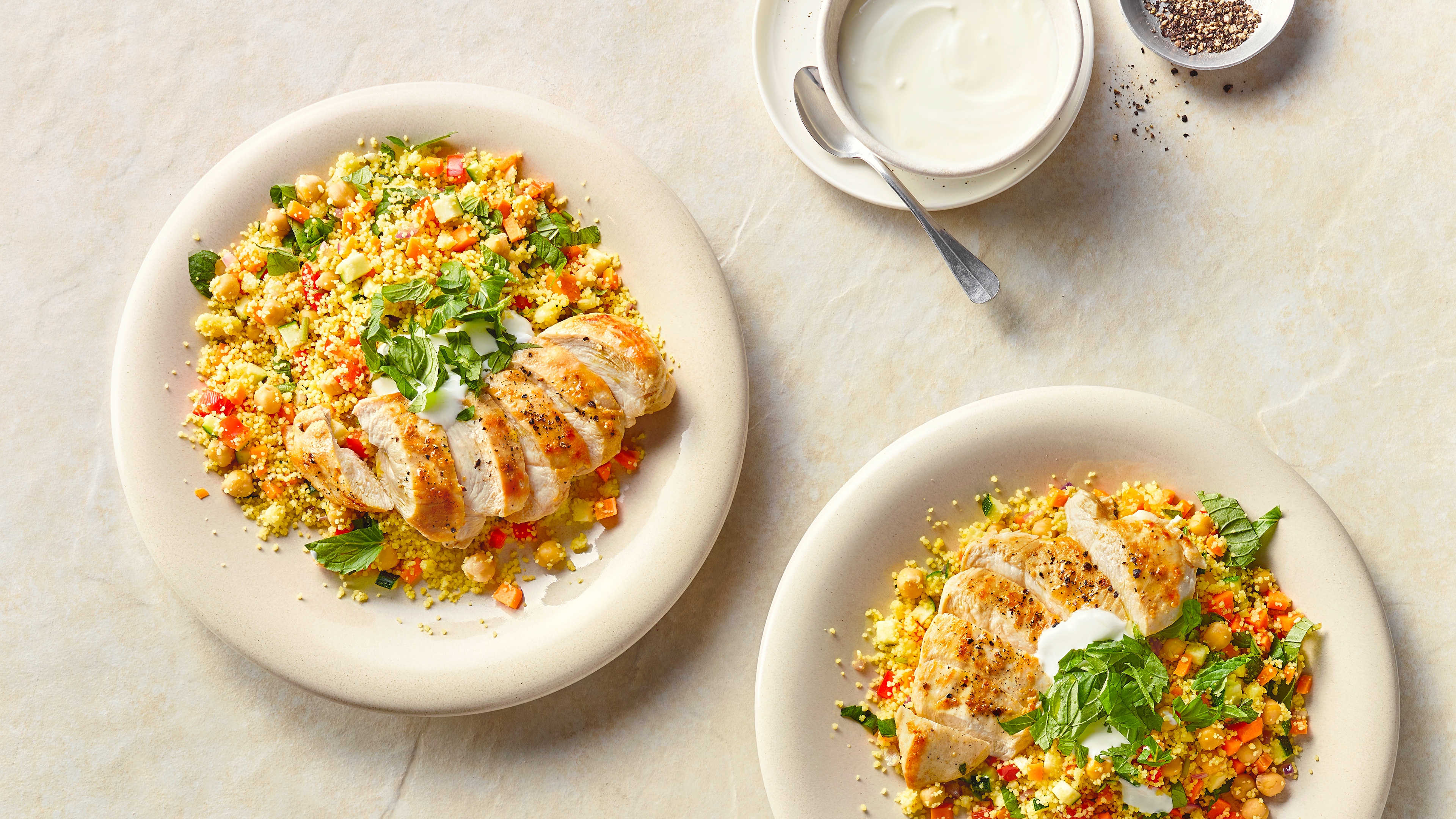 Ein Teller mit Couscous, Kichererbsen, Gurke, Paprika und Karotte, getoppt mit gebratenem Hähnchen und weißer Soße